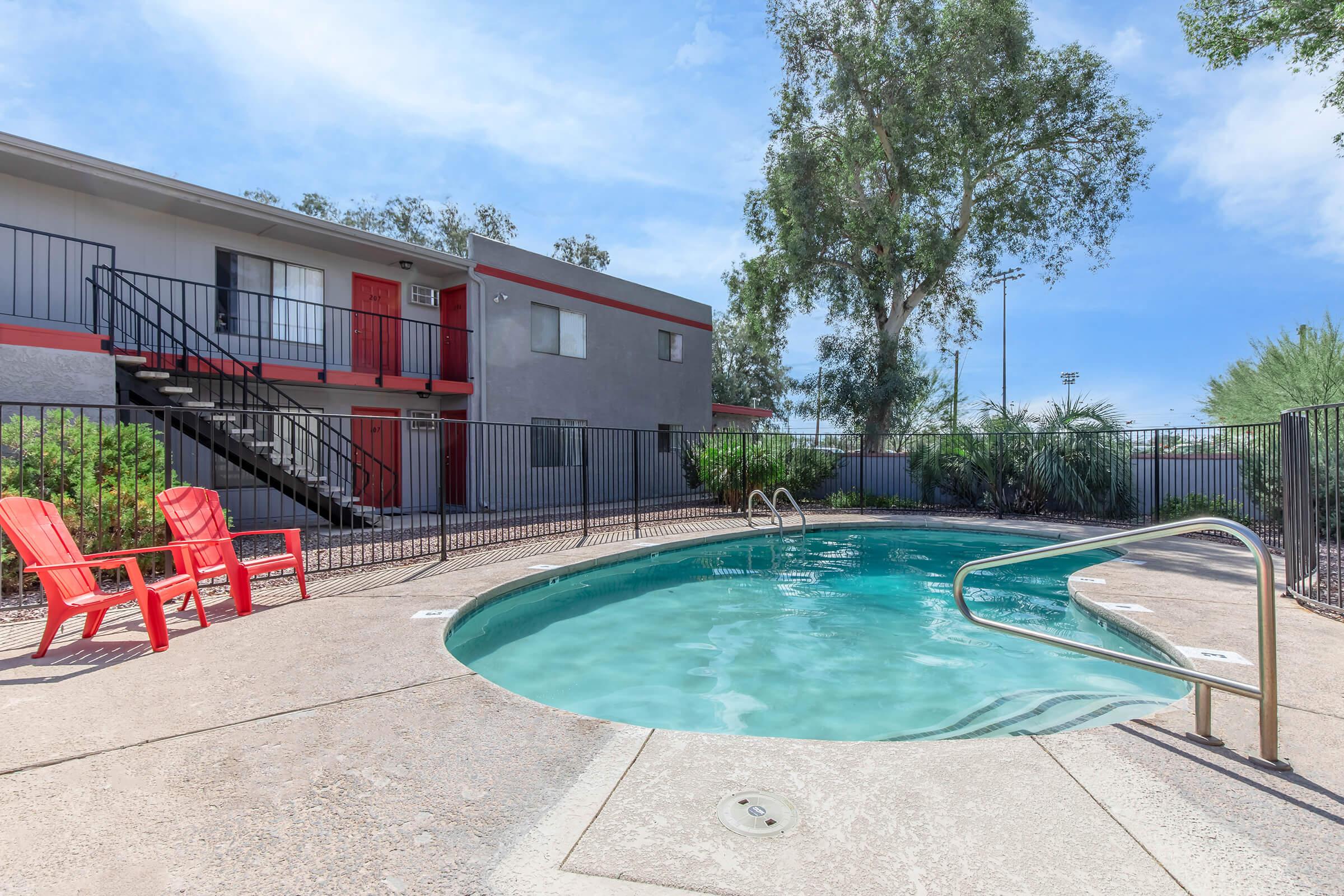 A swimming pool surrounded by a fence, featuring a set of red lounge chairs nearby. In the background, a two-story building with red doors and windows is visible, along with green trees and shrubs under a clear blue sky.