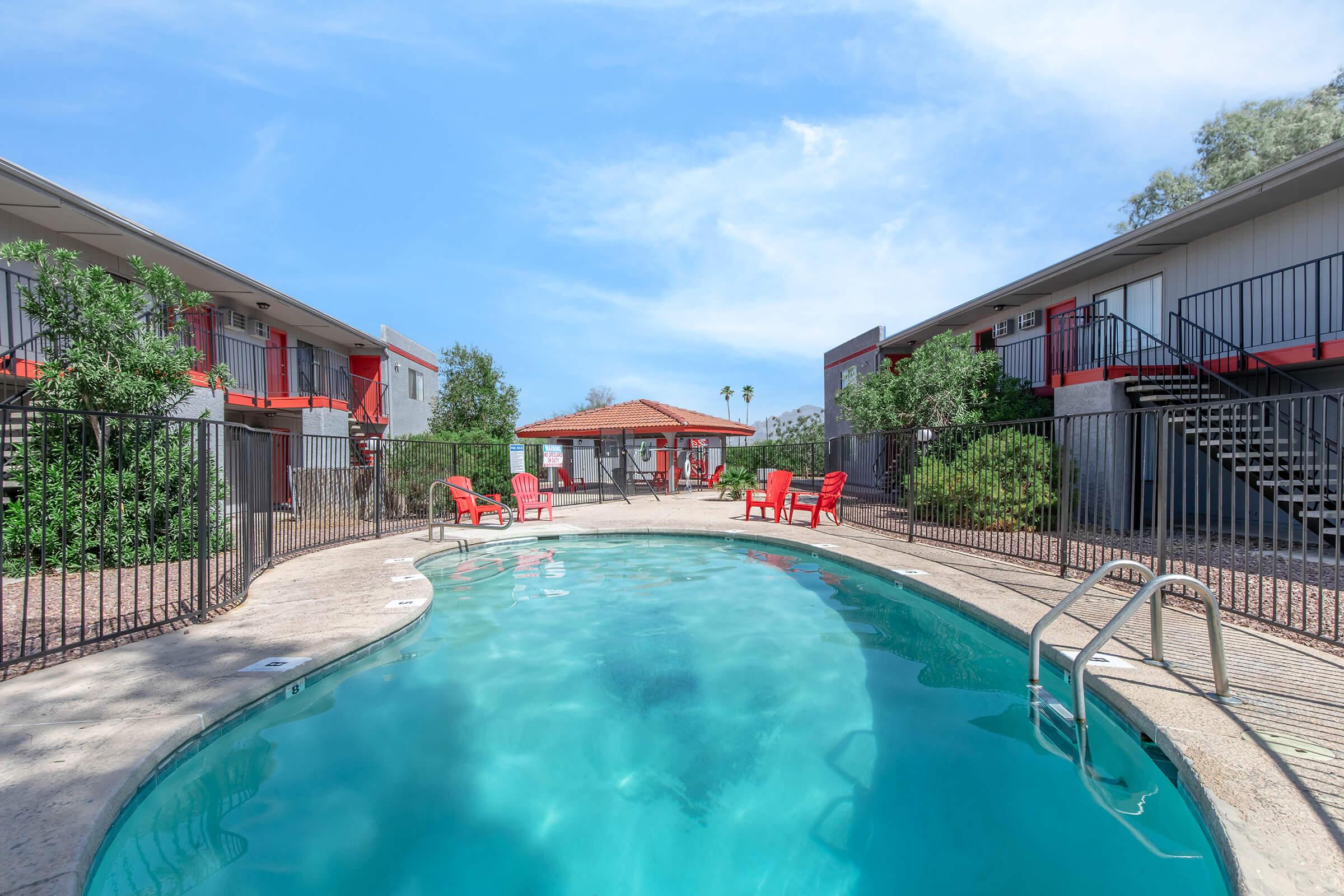 A clear swimming pool surrounded by a paved area, featuring red lounge chairs. In the background, there are two buildings with red accents and a gazebo-like structure. Lush greenery and palm trees are visible, creating a relaxing outdoor atmosphere under a blue sky.