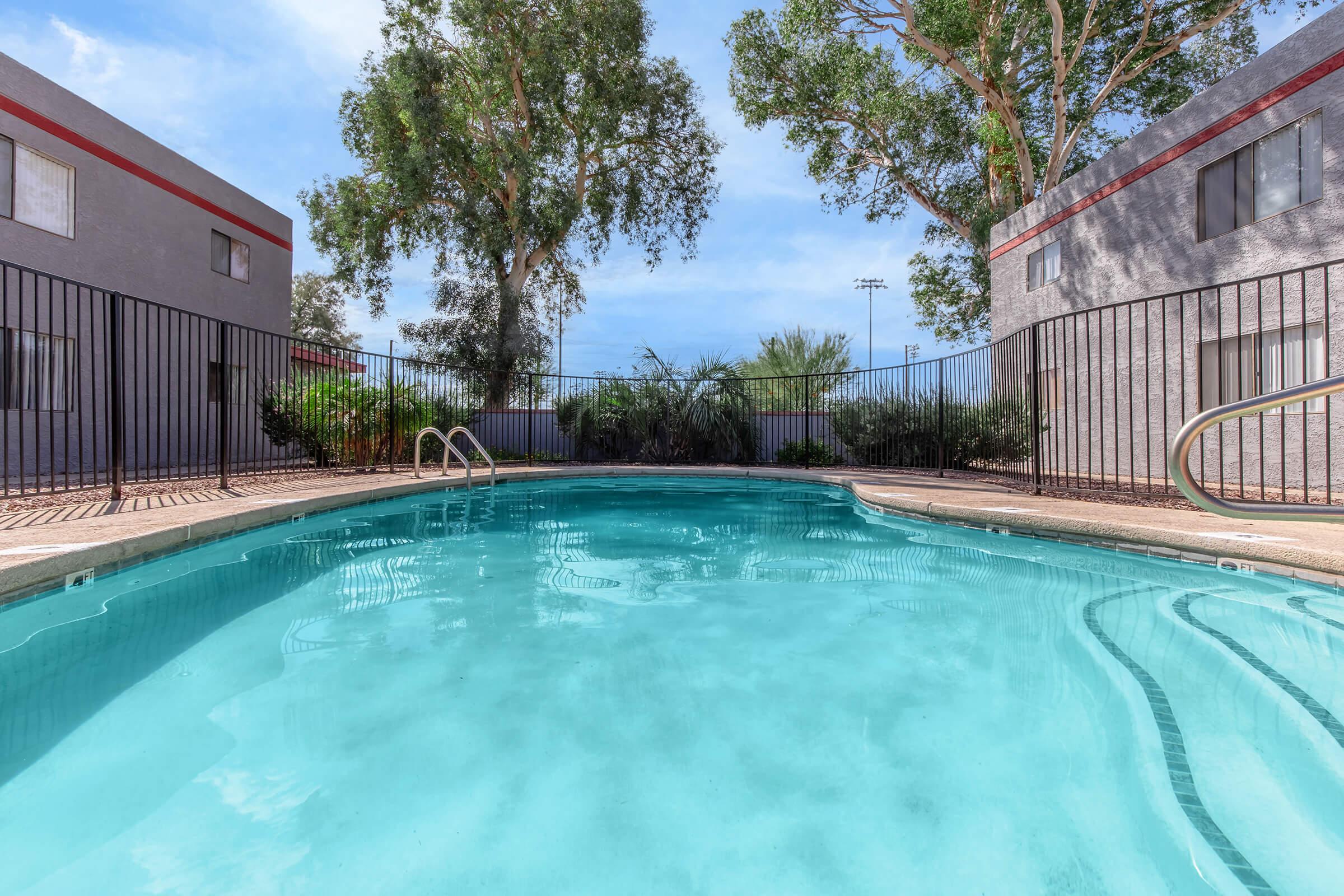 A serene swimming pool surrounded by a black wrought iron fence, situated between two gray apartment buildings. Lush greenery and trees are visible in the background, under a clear blue sky. The pool's water reflects the bright sunlight, creating an inviting atmosphere for relaxation and leisure.