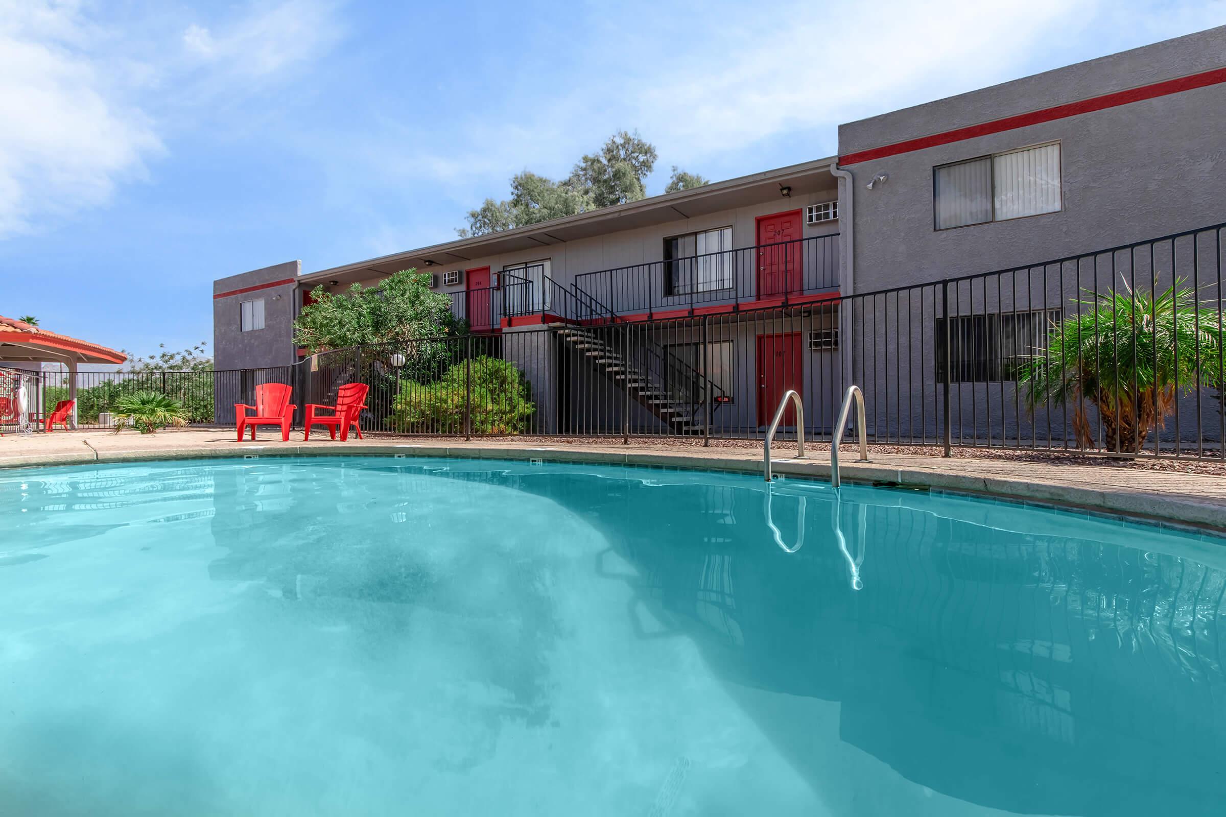 A clear swimming pool in the foreground with a surrounding black fence. Behind it, there is a two-story building featuring red doors and windows. Bright red lounge chairs are placed beside the pool, with palm trees and shrubs adding greenery to the scene. The sky is partly cloudy, enhancing a relaxed atmosphere.