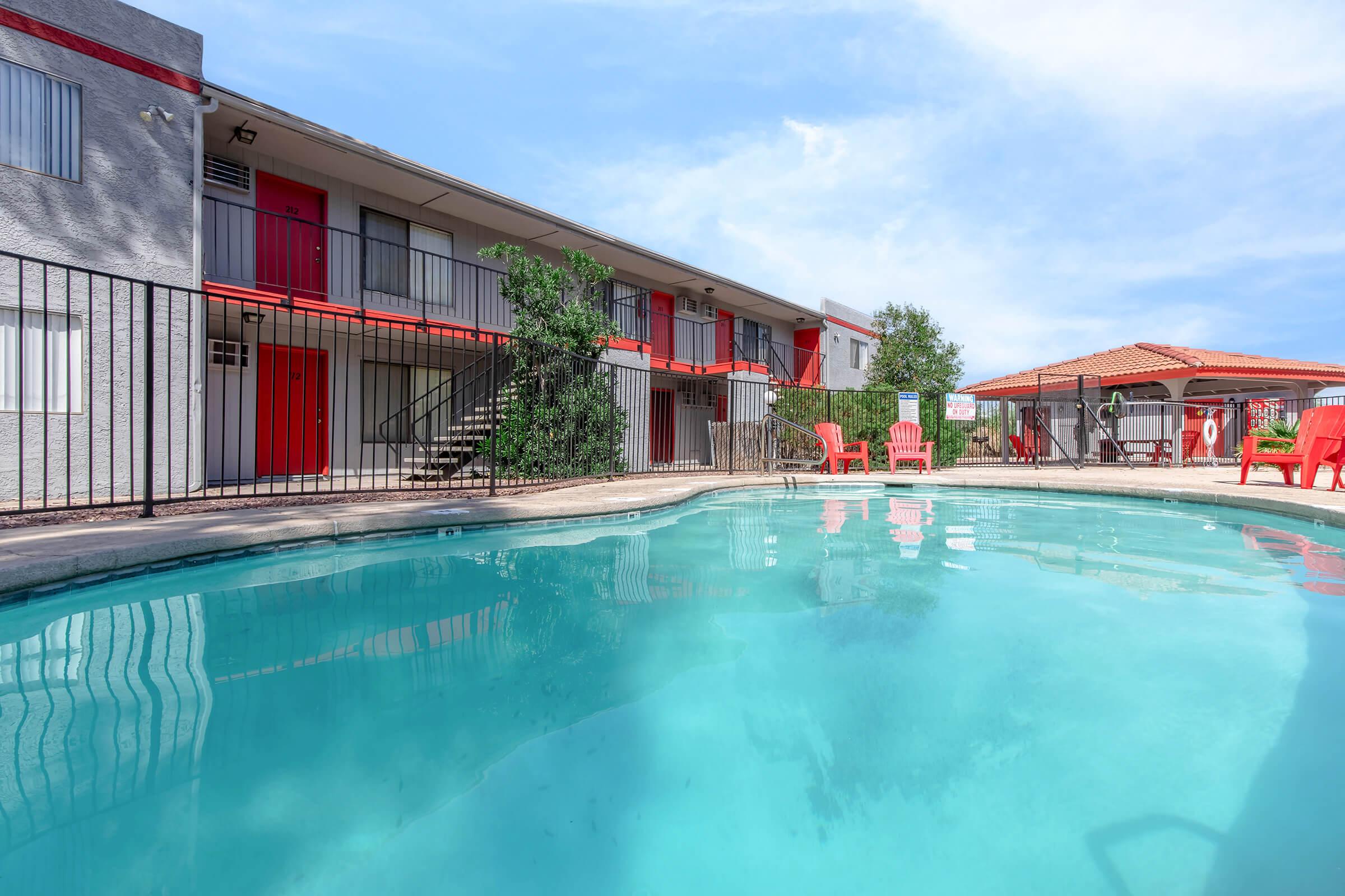 A swimming pool surrounded by outdoor seating, with red lounge chairs nearby. There's a two-story building in the background featuring balconies with red doors. A covered gazebo can be seen to the right, set against a clear blue sky with a few scattered clouds.