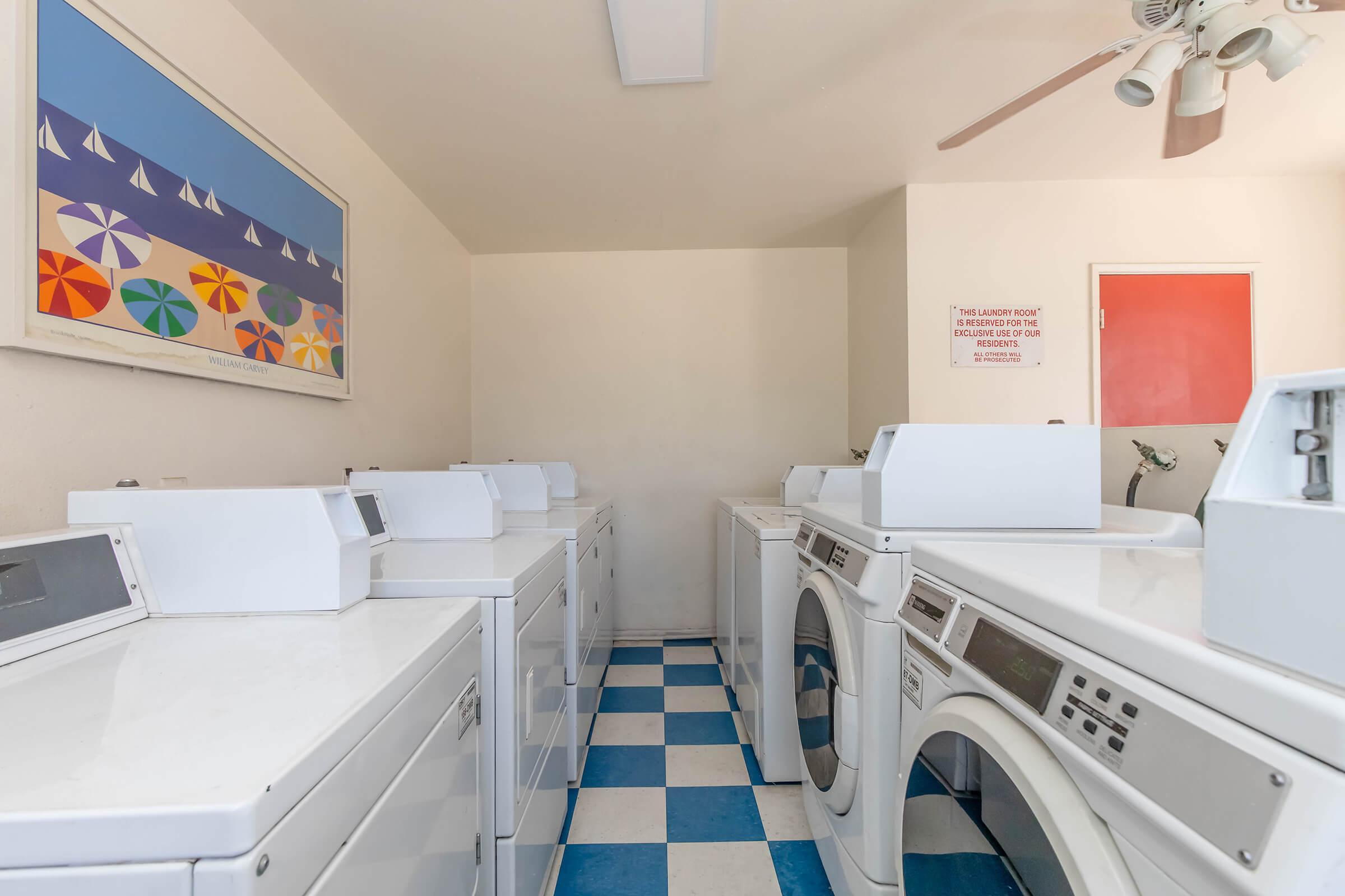 A clean laundry room featuring several white washing machines and dryers lined against the walls. The floor has a blue and white checkered pattern, and a colorful beach-themed artwork hangs on the wall. A ceiling fan is visible, and a bright red door is partially open at the far end of the room.