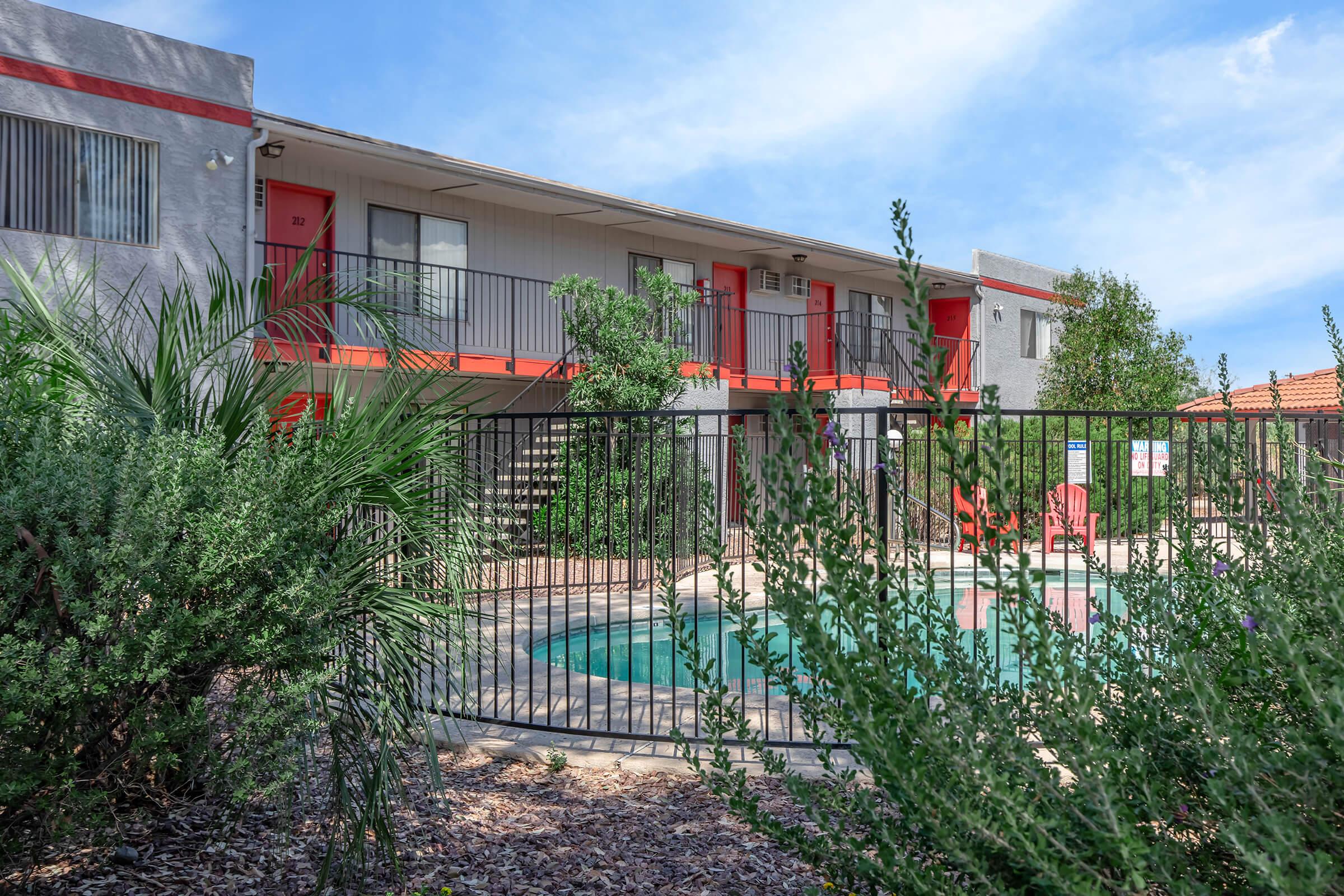 A view of an apartment building with a pool area, surrounded by greenery. The building features colorful red accents and balconies, with a staircase visible. The pool is enclosed by a fence, and there are lounge chairs nearby under a clear blue sky.