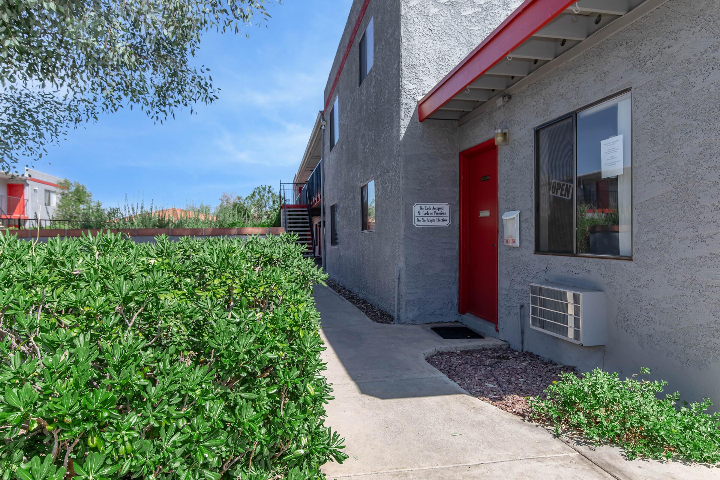 A walkway leading to a gray building with a red door and window. Lush green bushes are in the foreground, and there are steps leading to the upper level of the building. Clear blue sky is visible above, indicating a sunny day.