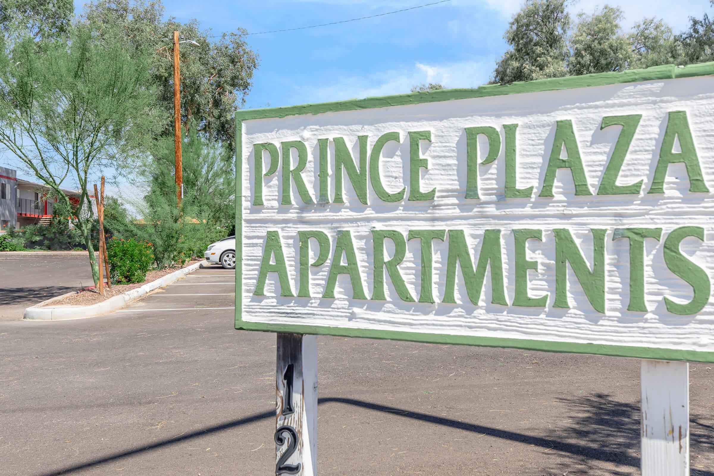 Sign for Prince Plaza Apartments, with a textured white background and green lettering. Surrounding area features trees and a parking lot with vehicles. Blue sky and a few clouds are visible in the background.
