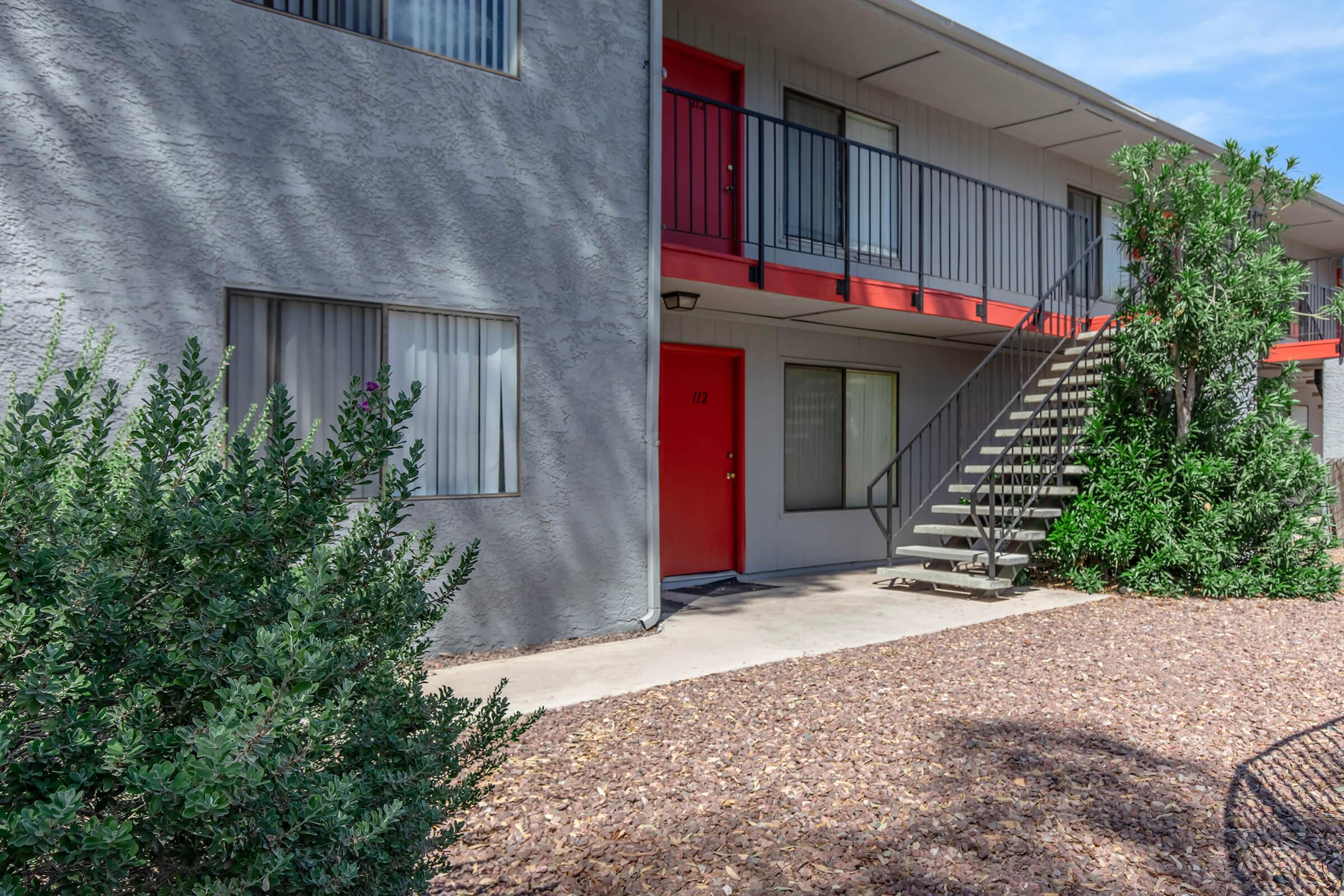 Exterior view of a two-story apartment building with a gray façade and red doors. A staircase leads to the upper level. The ground is covered with small pebbles, and there are shrubs in the foreground. Large windows allow natural light in, and the scene is set under a clear blue sky.