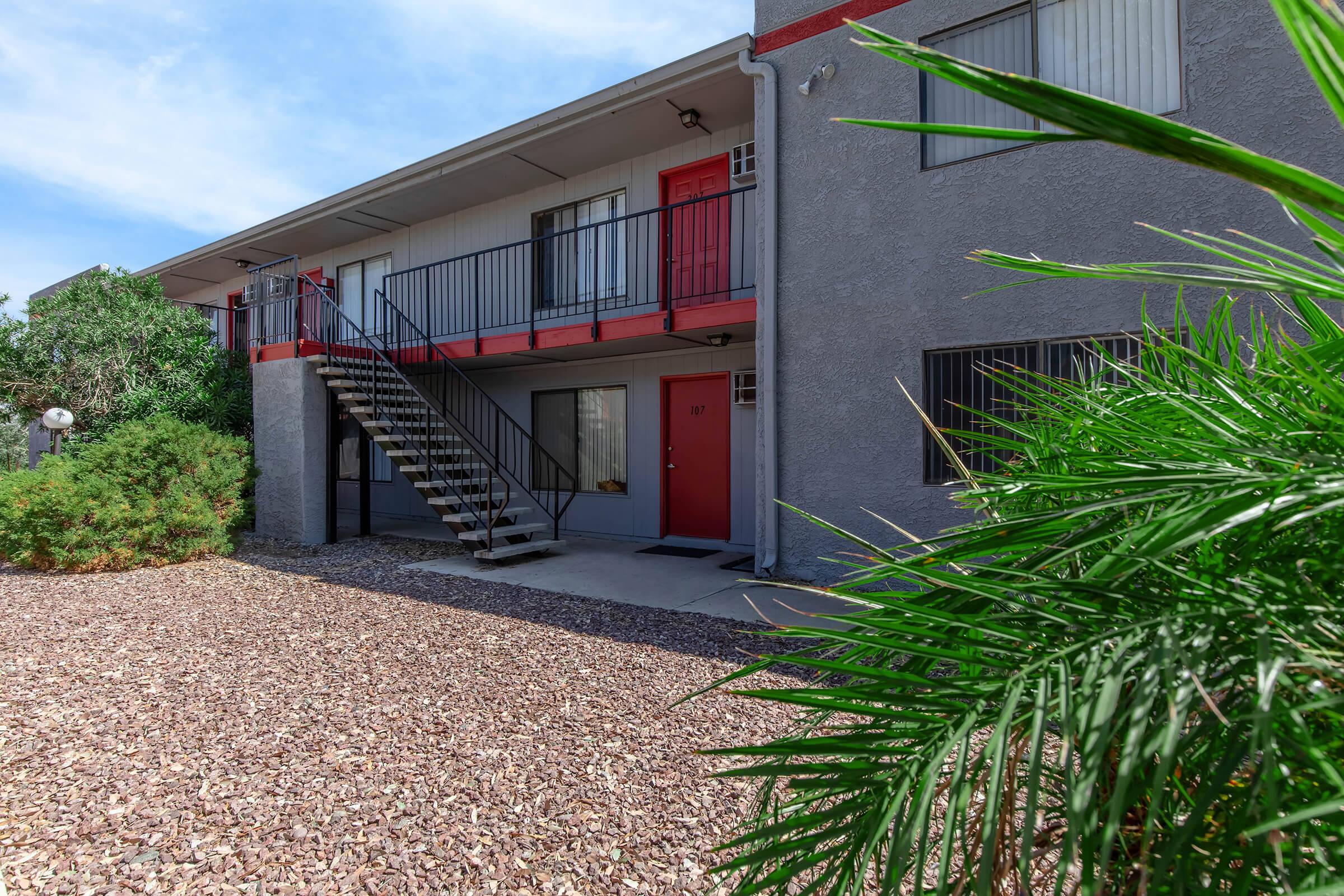 A view of a two-story apartment building featuring a staircase leading to the second floor. The building has a red door on the bottom level, surrounded by landscaping with low shrubs and rocks. The sky is clear with some clouds, indicating a bright day.