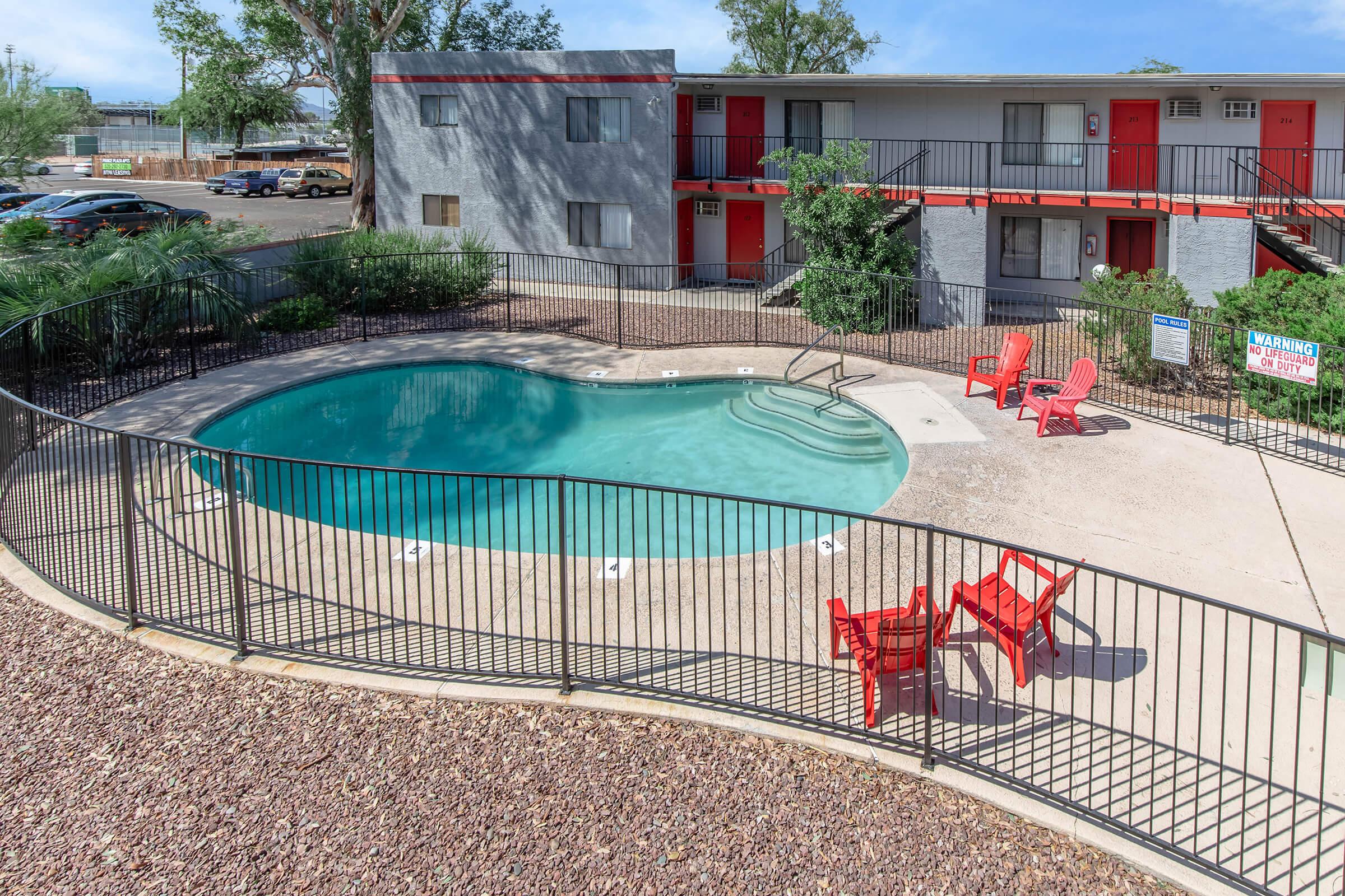 A view of a swimming pool surrounded by a black fence, featuring red patio chairs on one side. In the background, there are two-story apartment buildings with red doors and a landscaped area with shrubs and trees. The scene is bright and inviting, ideal for relaxation.