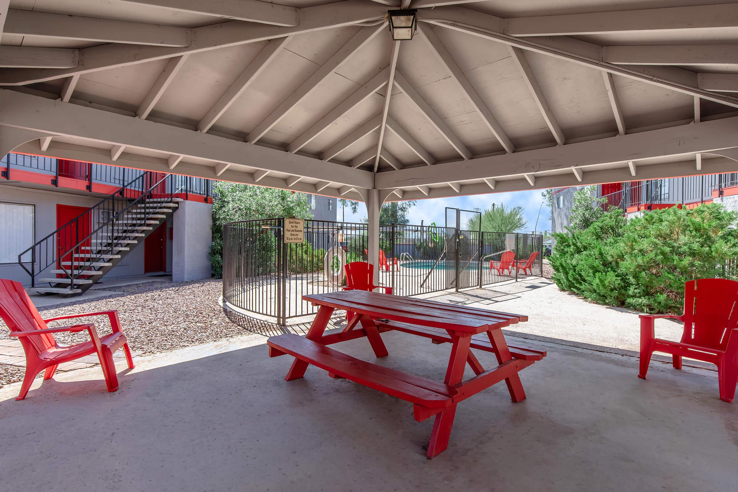 A shaded pavilion with a red picnic table and chairs, surrounded by greenery. In the background, a pool area is visible, enclosed by a fence. A staircase leads to upper levels of nearby buildings, which are painted in a contrasting color. The scene is well-lit with natural sunlight.