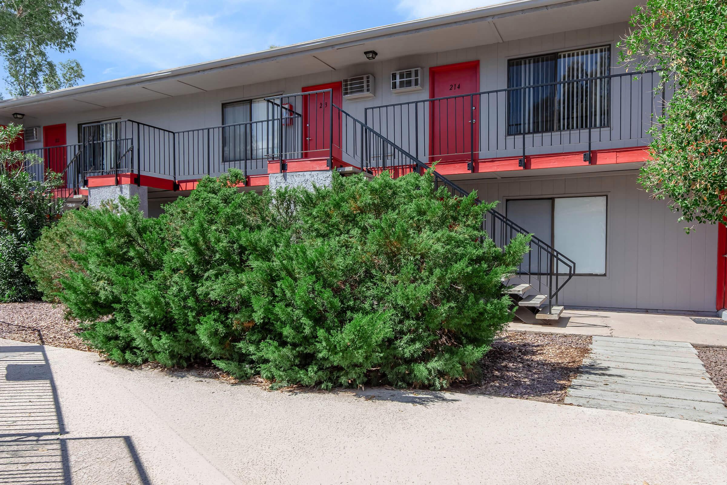 A view of a two-story apartment building with red doors and black railings. The building is surrounded by greenery and a pathway, with a well-maintained landscape featuring gravel and shrubs. There are stairs leading to the upper level. Bright blue sky is visible above.