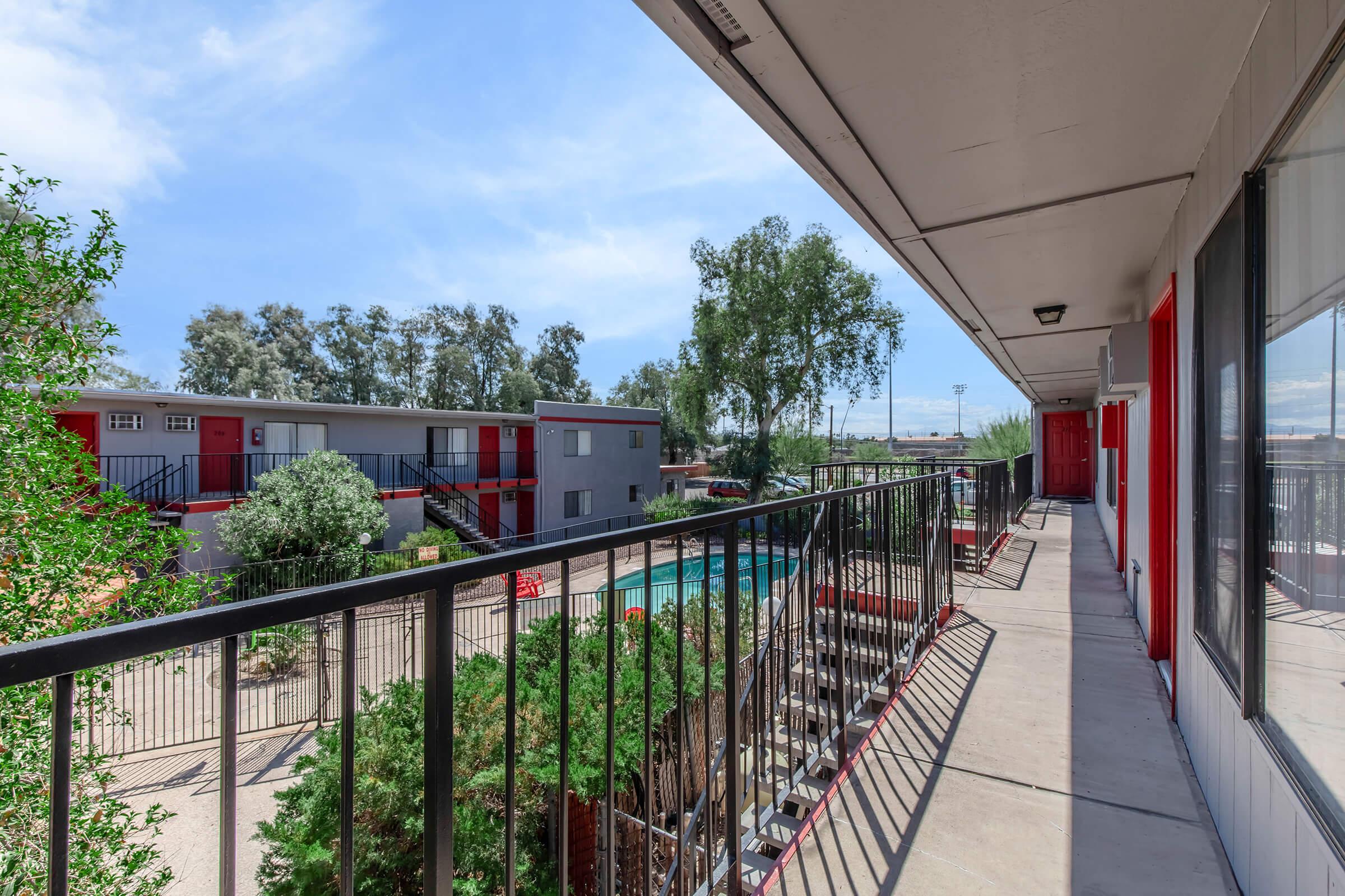 View from a second-floor balcony overlooking a courtyard with a pool. The scene features modern hotel-style buildings, greenery, and a clear blue sky. The balcony has a black railing, and the surroundings include trees and seating areas near the pool.