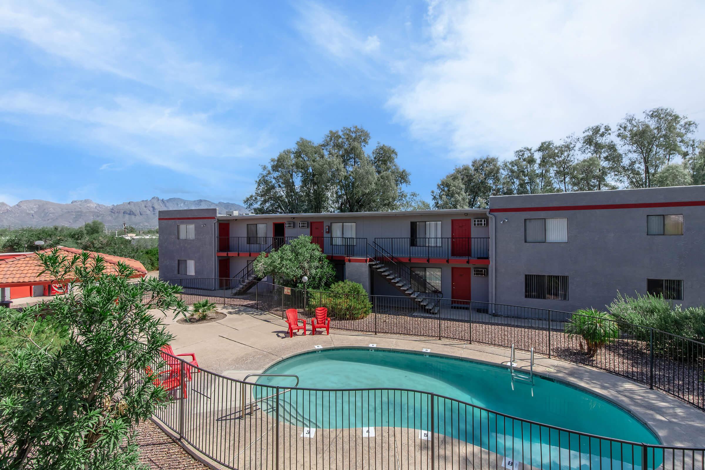A view of an apartment complex featuring a curved swimming pool surrounded by greenery. The two-story building has gray walls with red accents and stairs leading to second-floor units. Bright red chairs are placed near the pool area, set against a backdrop of mountains and a blue sky.