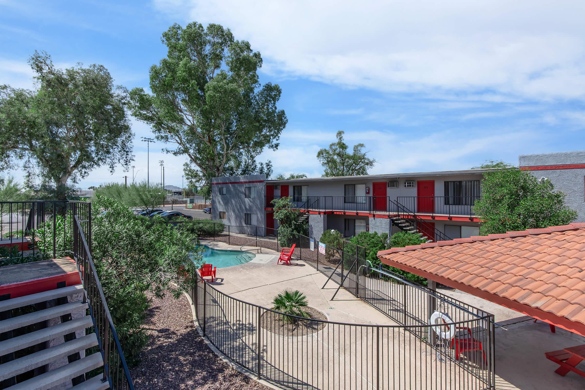 View of a courtyard in an apartment complex featuring a swimming pool surrounded by green trees and red lounge chairs. The two-story building has red doors and balconies, with a staircase leading to the upper level. A shaded area with a tiled roof is visible nearby.
