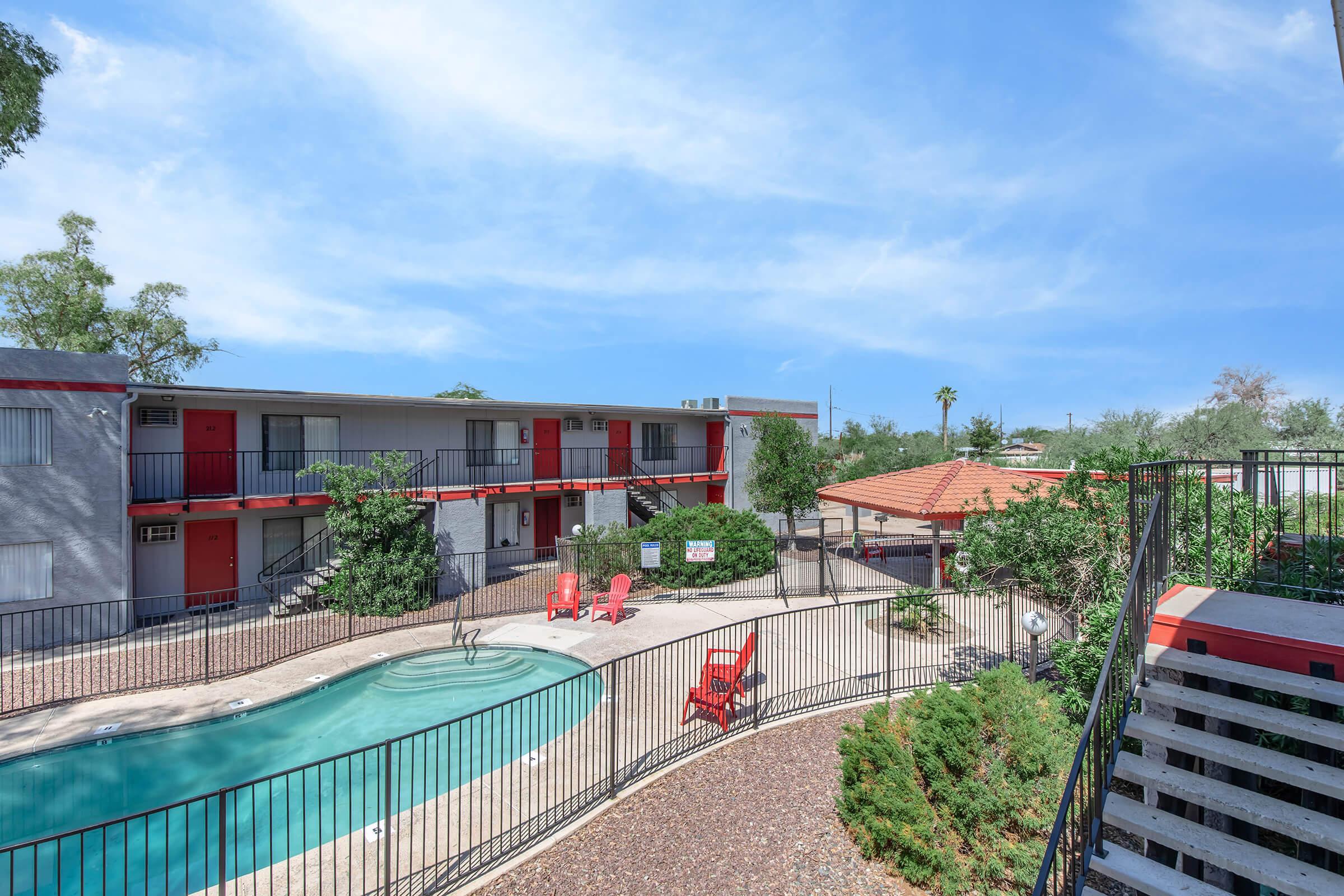 View of a modern apartment complex featuring a swimming pool surrounded by red lounge chairs, landscaped greenery, and a gazebo. The two-story buildings have red accents and are set against a clear blue sky, creating a welcoming atmosphere for residents.