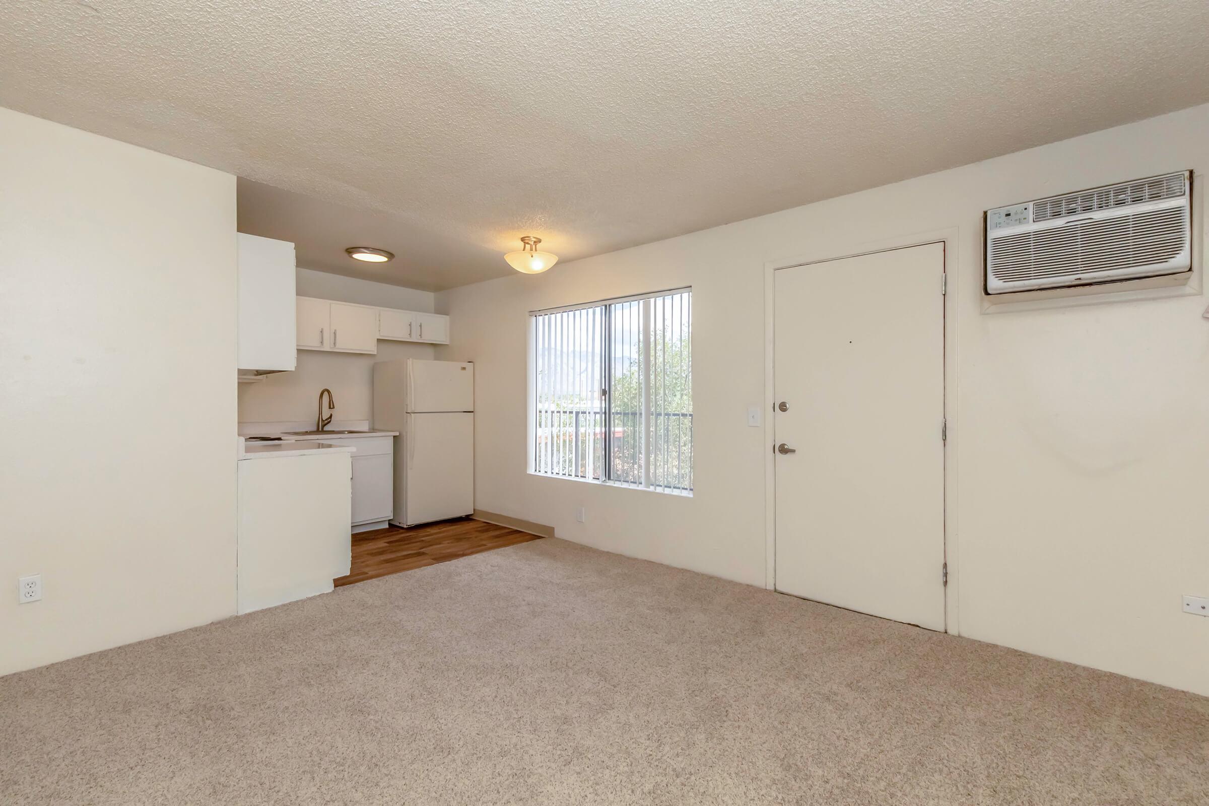 A cozy, unfurnished room with beige carpet, featuring a small kitchen area with white cabinets, a sink, and a refrigerator. A window lets in natural light, and there's a door leading outside. An air conditioning unit is mounted on the wall. The overall atmosphere is bright and inviting, perfect for personal decoration.