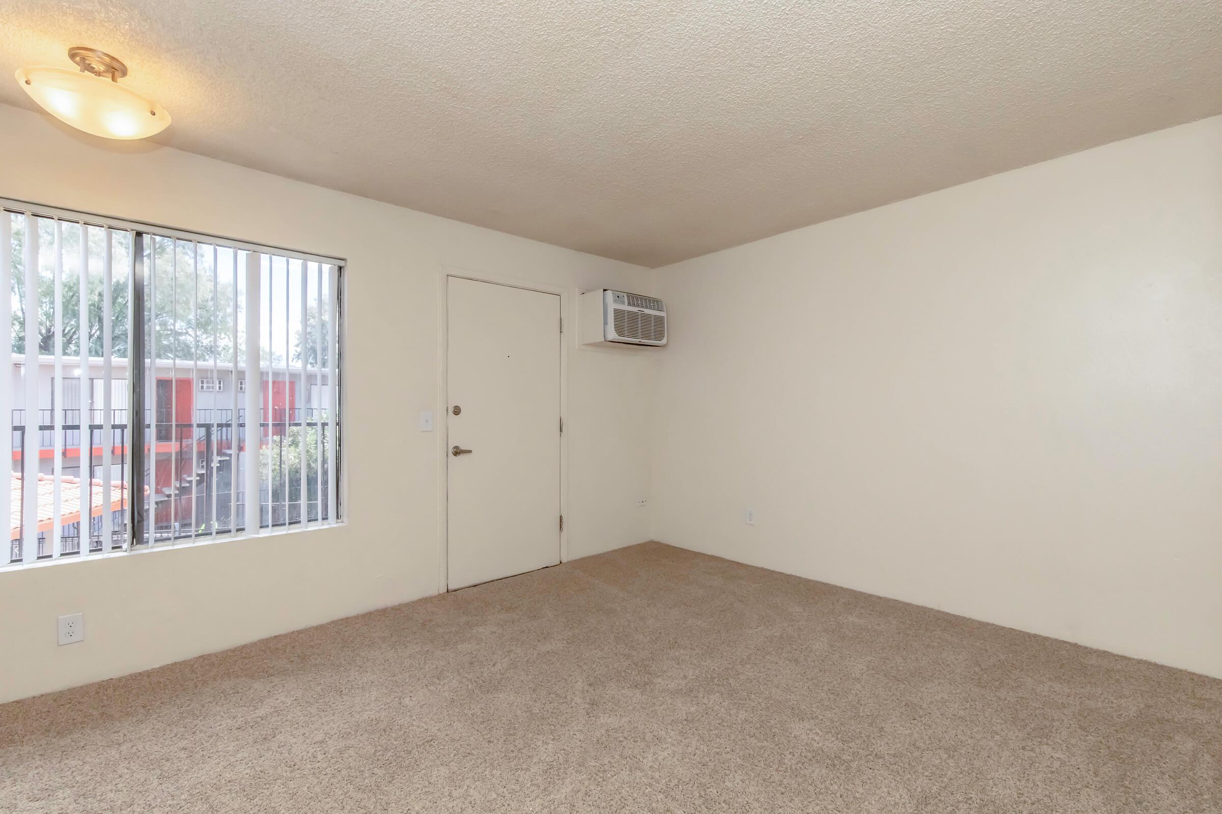 Empty room with beige carpet and light-colored walls. A door is on the left side, and a window with blinds is on the right side, allowing natural light. A small air conditioning unit is mounted above the door. The space appears clean and ready for furnishings.