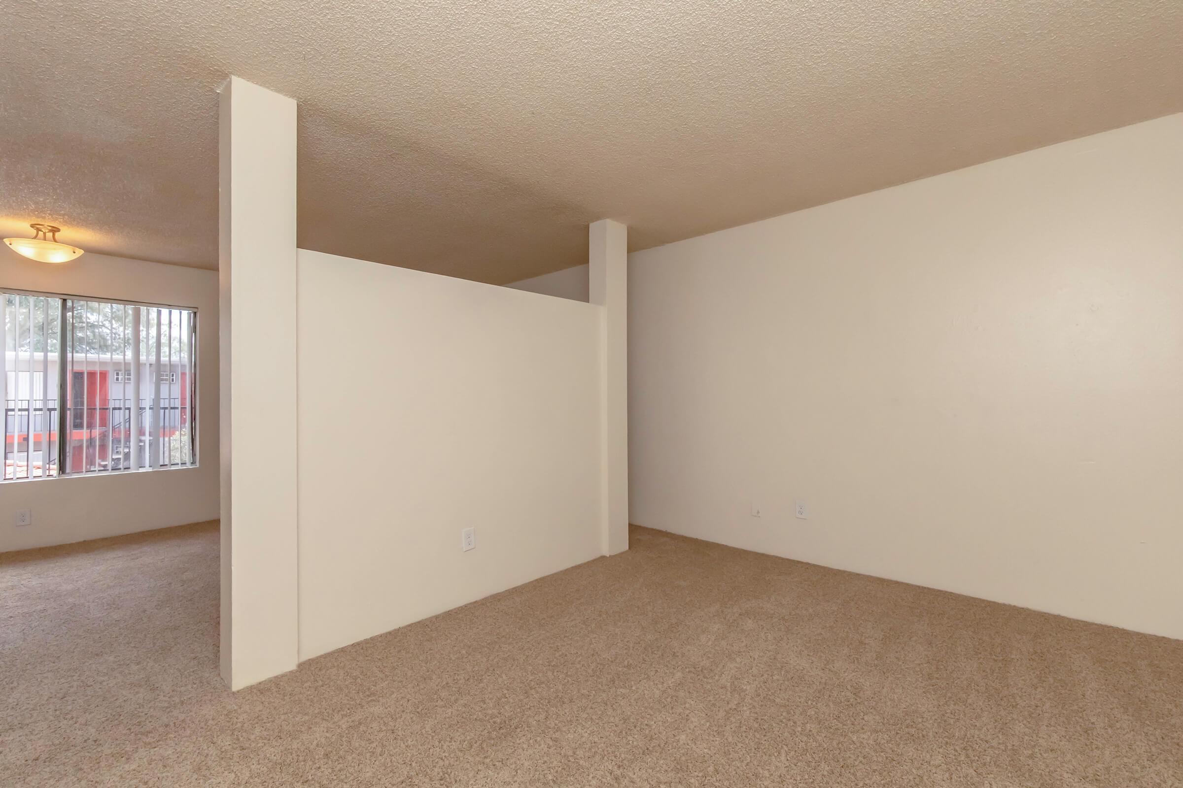 A spacious and empty room with beige carpet flooring and light-colored walls. There is a partition separating two areas, and a window with white blinds allows natural light to enter. The overall atmosphere is clean and inviting, suitable for personal decoration.