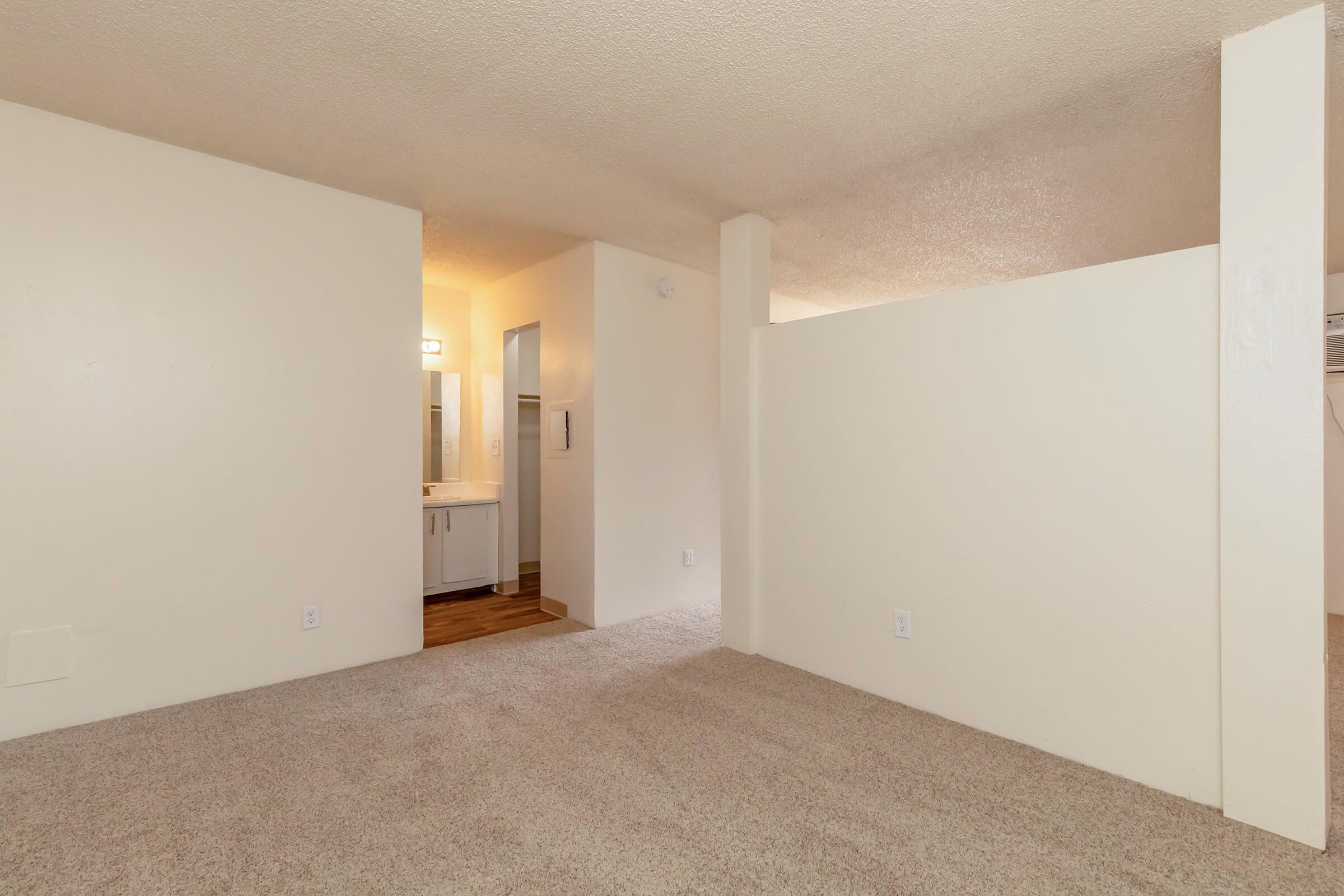 Interior view of a room showing beige carpet flooring and light-colored walls. A partial wall divides the space, with an entrance to a bathroom visible in the background. The lighting is soft, creating a warm atmosphere, and there are no furnishings in the room.