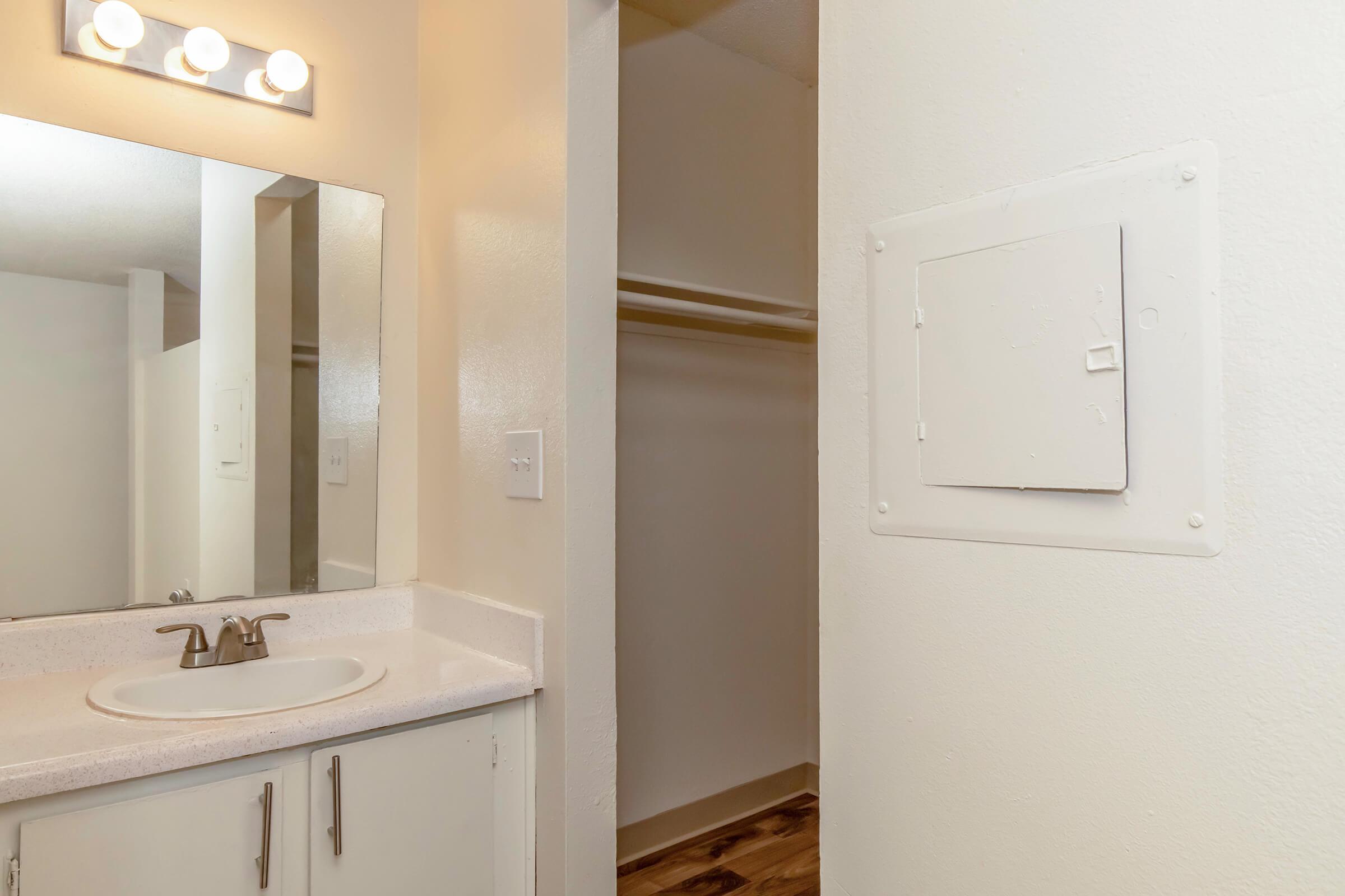 A small bathroom featuring a single sink with a mirror above it. The sink is set against a light-colored countertop and cabinetry. To the right, there is an opening leading to a closet space, with a white wall and a small access panel. The room has neutral-colored walls and a warm-toned floor.
