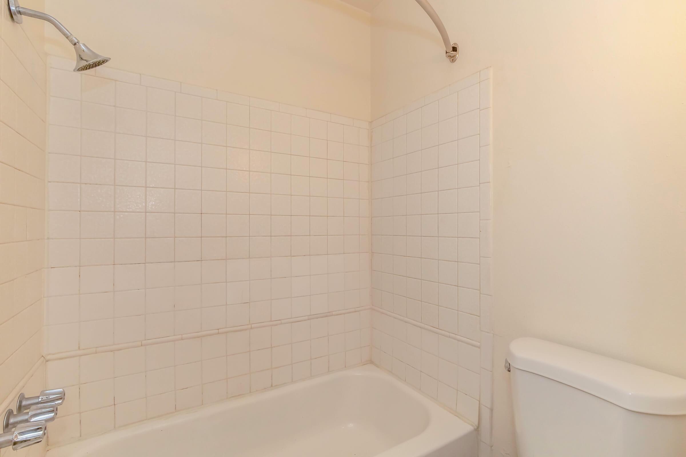 A bathroom featuring a white tiled shower area with a bathtub and a toilet. The walls are plain and light-colored, and the showerhead is mounted on the wall above the tub. The overall appearance is clean and minimalistic.