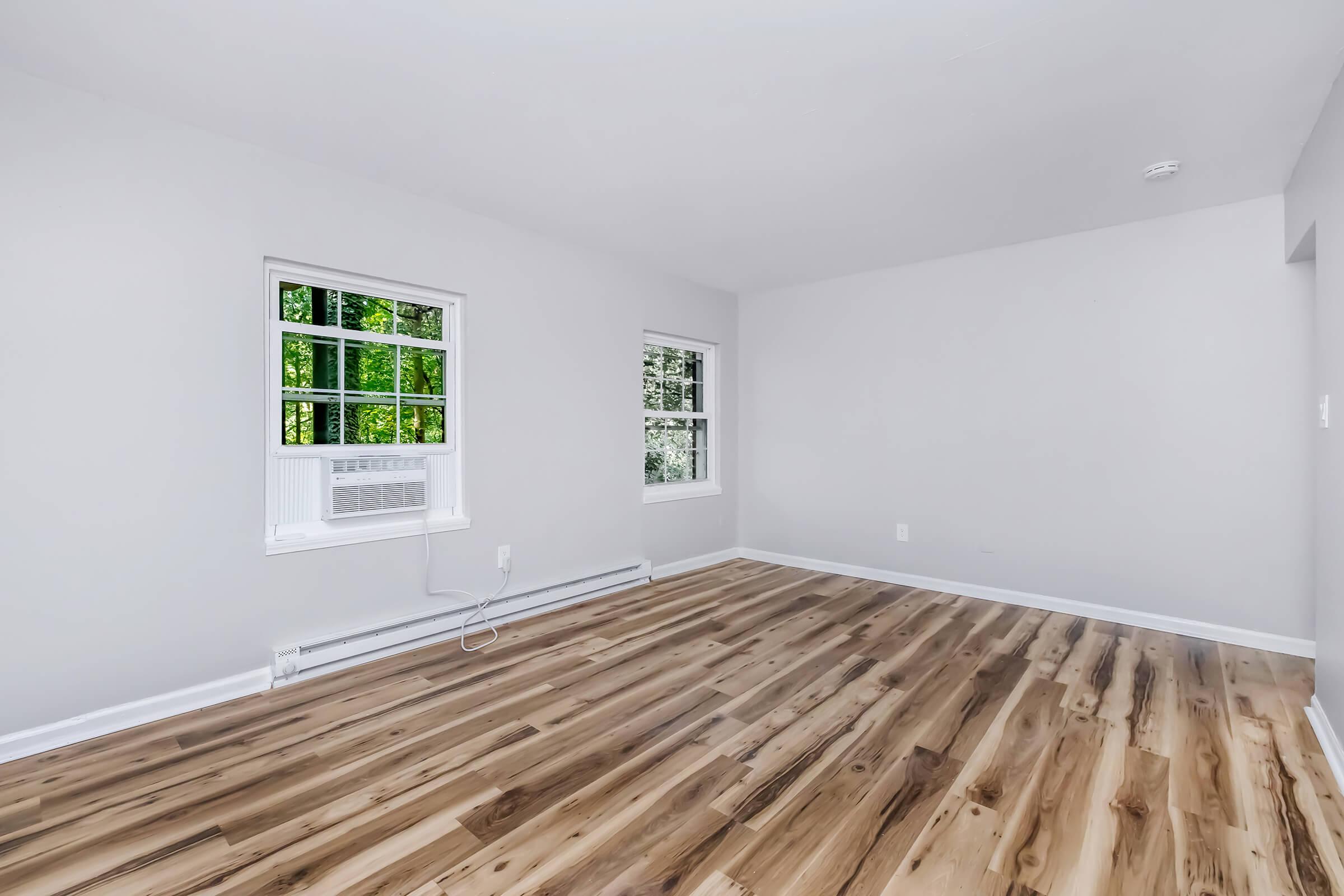A bright, empty room with light grey walls and hardwood floors. Two windows allow natural light to fill the space, and there is a window air conditioning unit on one wall. The overall atmosphere is clean and modern, suitable for various interior design options.