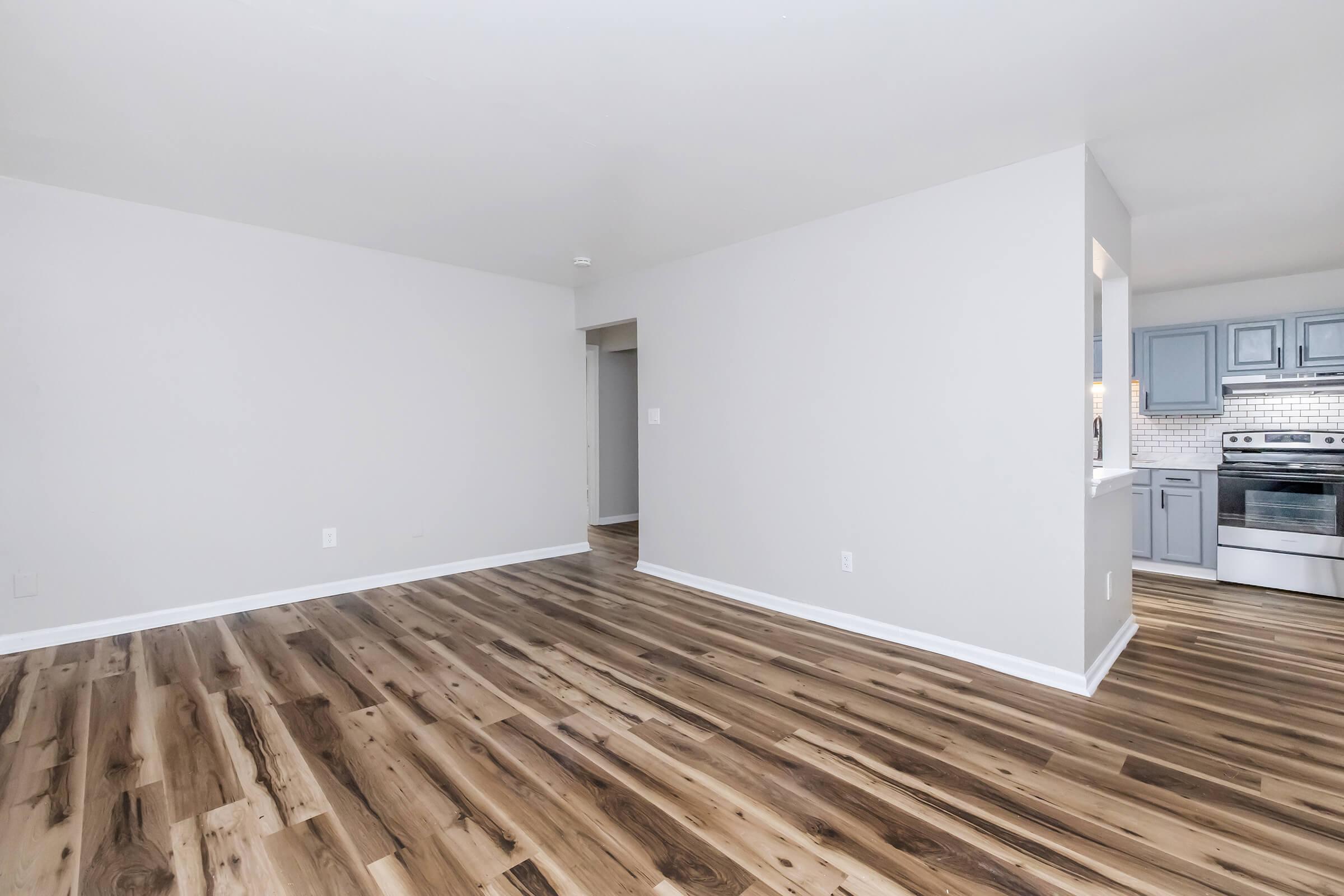 A spacious, unfurnished living area with light-colored walls and modern wood flooring. On the right, there's a glimpse of a kitchen with gray cabinetry and stainless steel appliances. The area is well-lit, creating an inviting atmosphere.