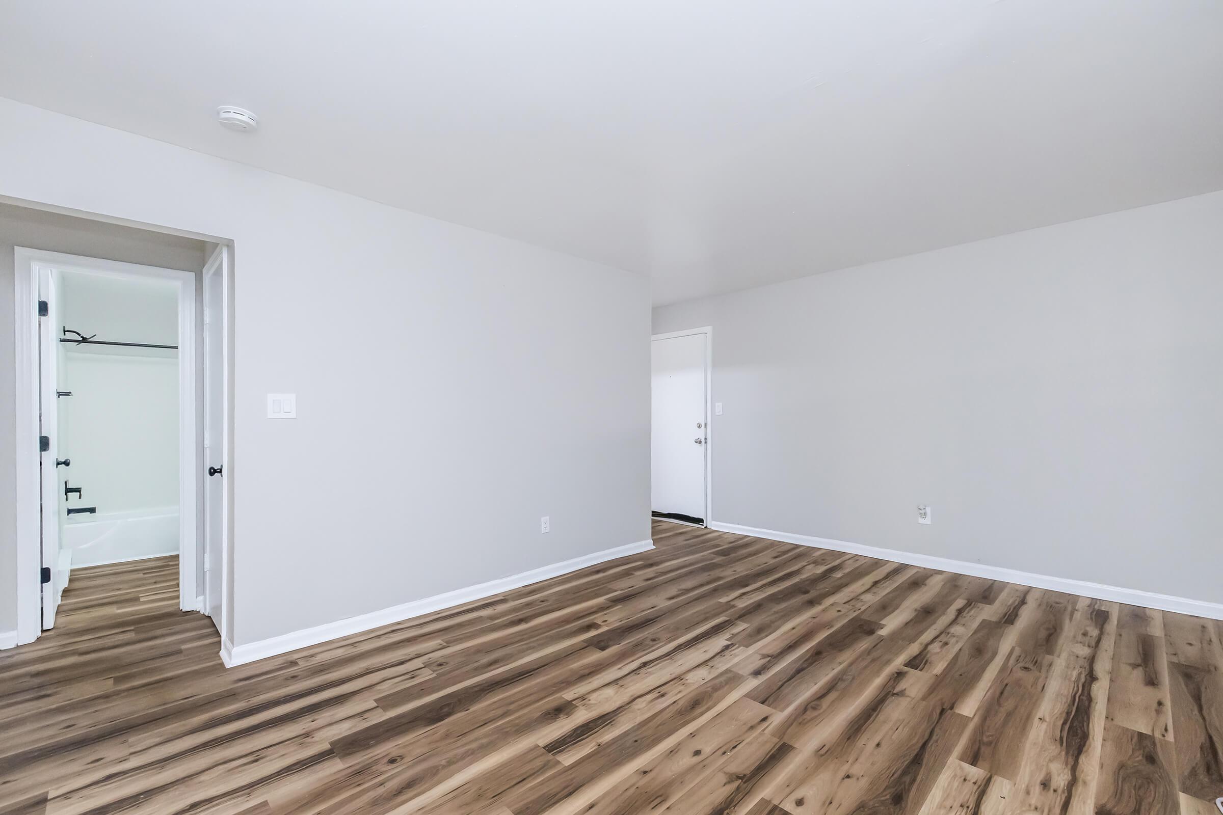 A spacious, empty room with light gray walls and hardwood flooring. There are two doorways visible: one leads to a bathroom with a tub and the other leads to another part of the living space. The room has ample natural light and appears freshly painted and newly renovated.