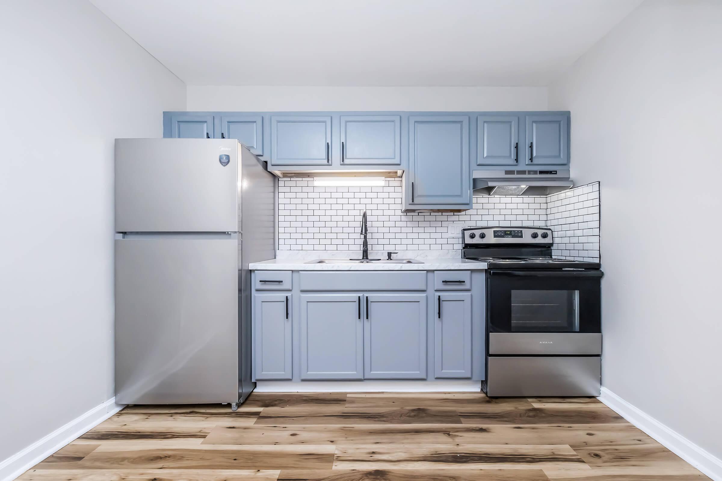 A modern kitchen featuring light blue cabinets, a stainless steel refrigerator, an oven, and a white subway tile backsplash. The space has a neutral wall color and warm wood flooring, creating a bright and inviting atmosphere.