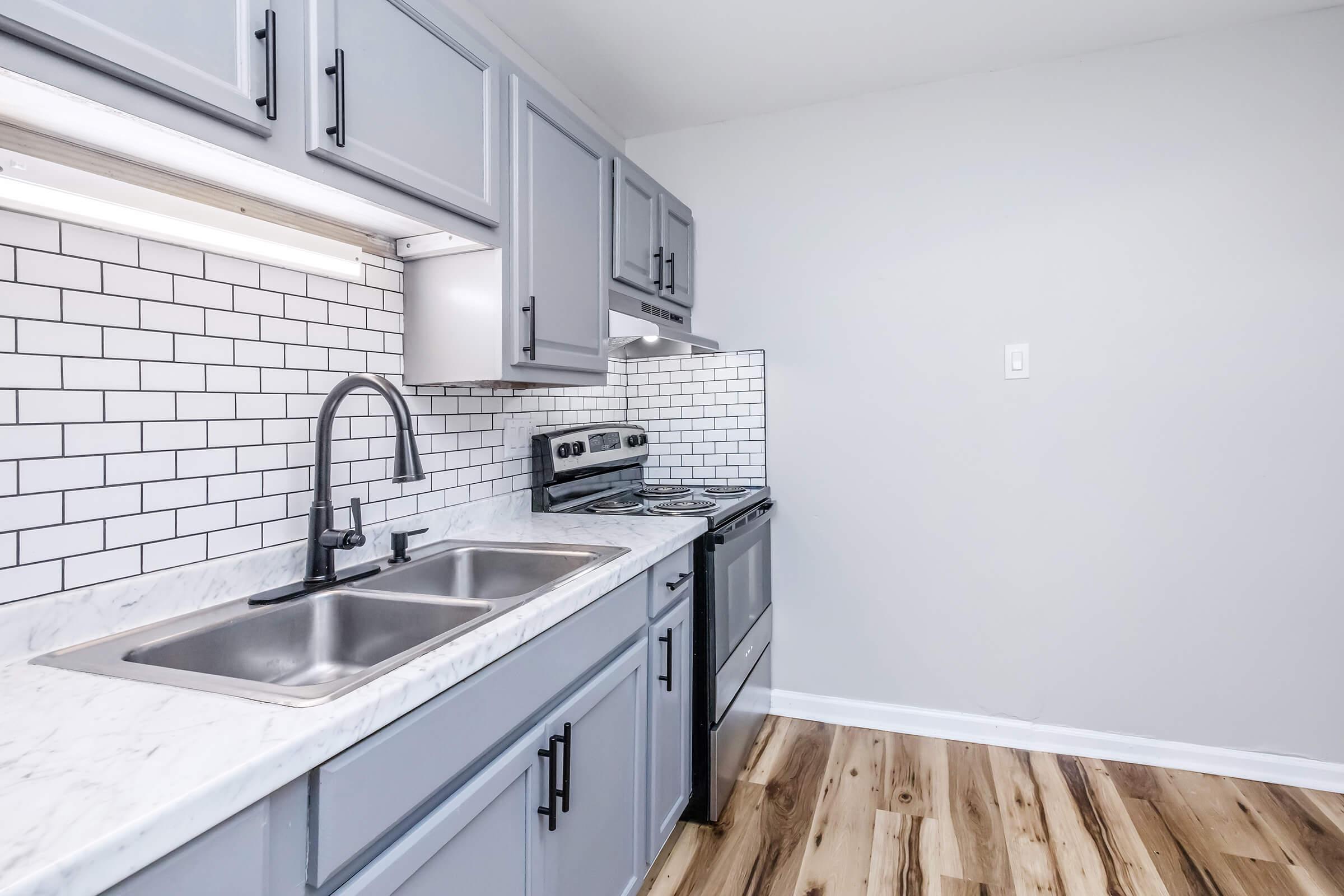 A modern kitchen featuring gray cabinetry, a double sink with a sleek faucet, a stainless steel stove, and a white subway tile backsplash. The countertop is marble, and the flooring showcases light wood tones, creating a bright and inviting space.