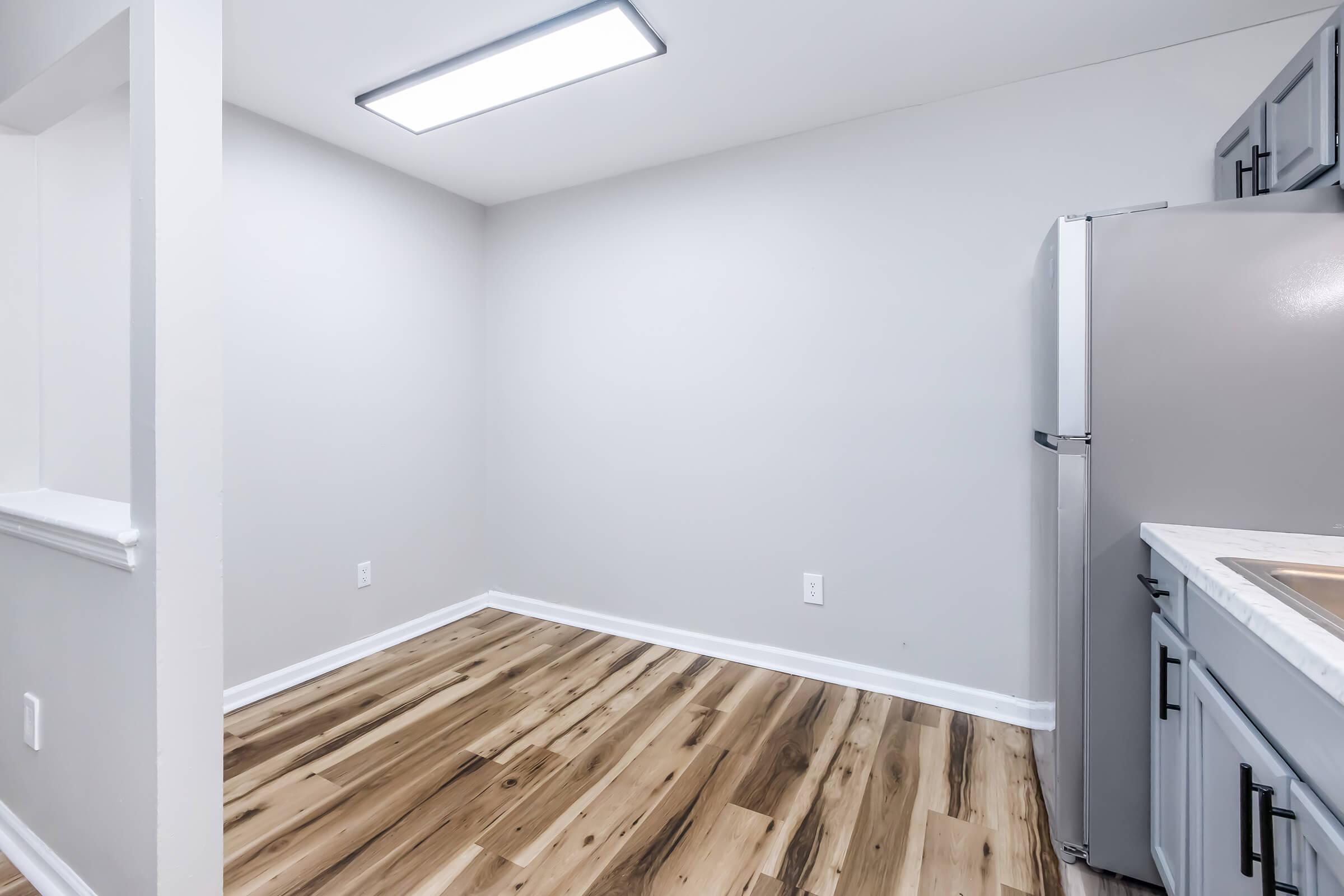 A modern kitchen with light gray walls and a wooden floor. The image features a stainless steel refrigerator on the right and a countertop with a sink on the right side. The ceiling has a bright overhead light, and the space appears clean and empty, ready for furniture or appliances.