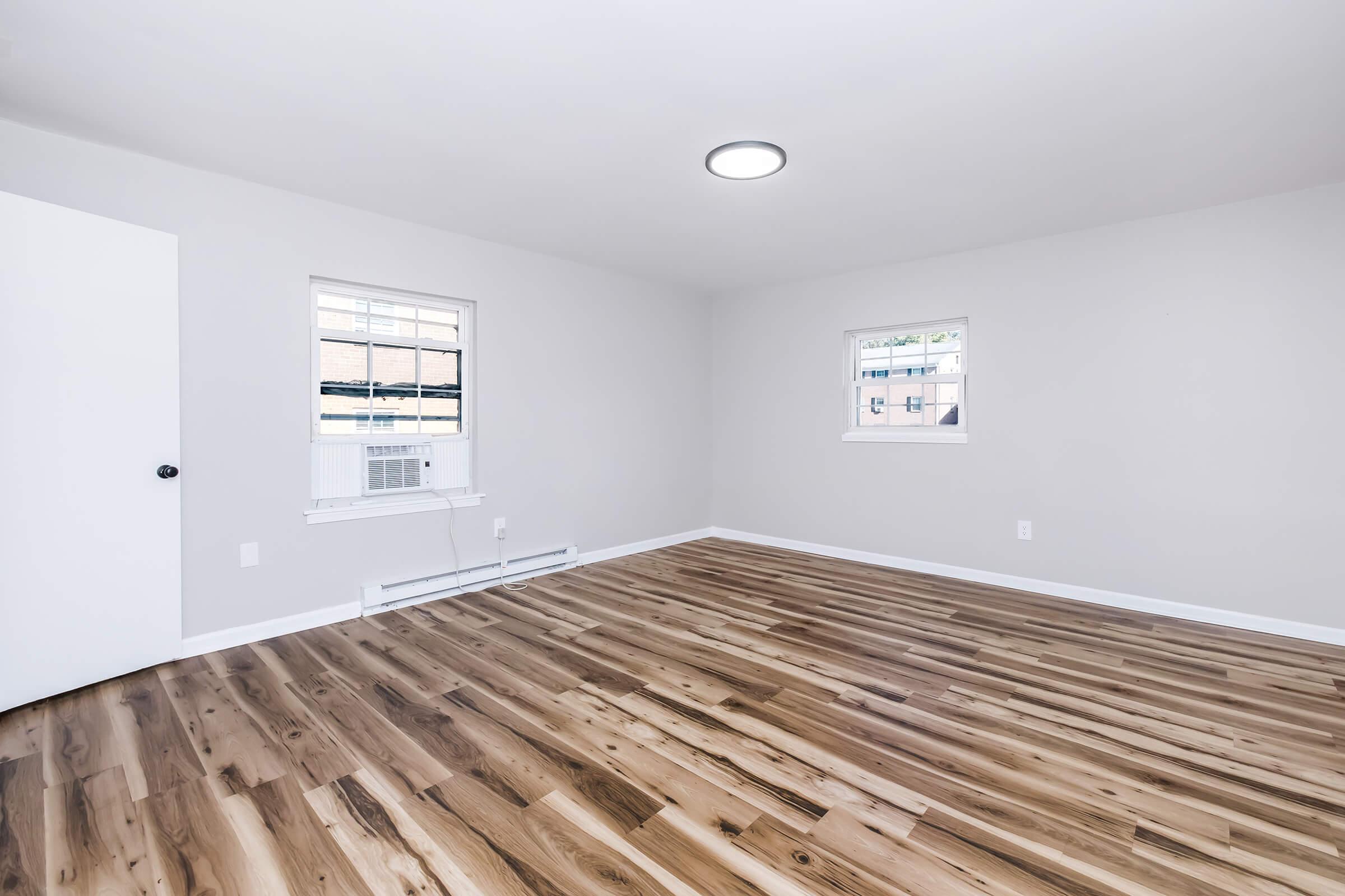 Empty room with light gray walls and hardwood flooring. Two small windows allow natural light, and there is a door on the left side. An air conditioning unit is visible beneath one window. The ceiling has a modern round light fixture. The space appears clean and freshly painted, suitable for various uses.