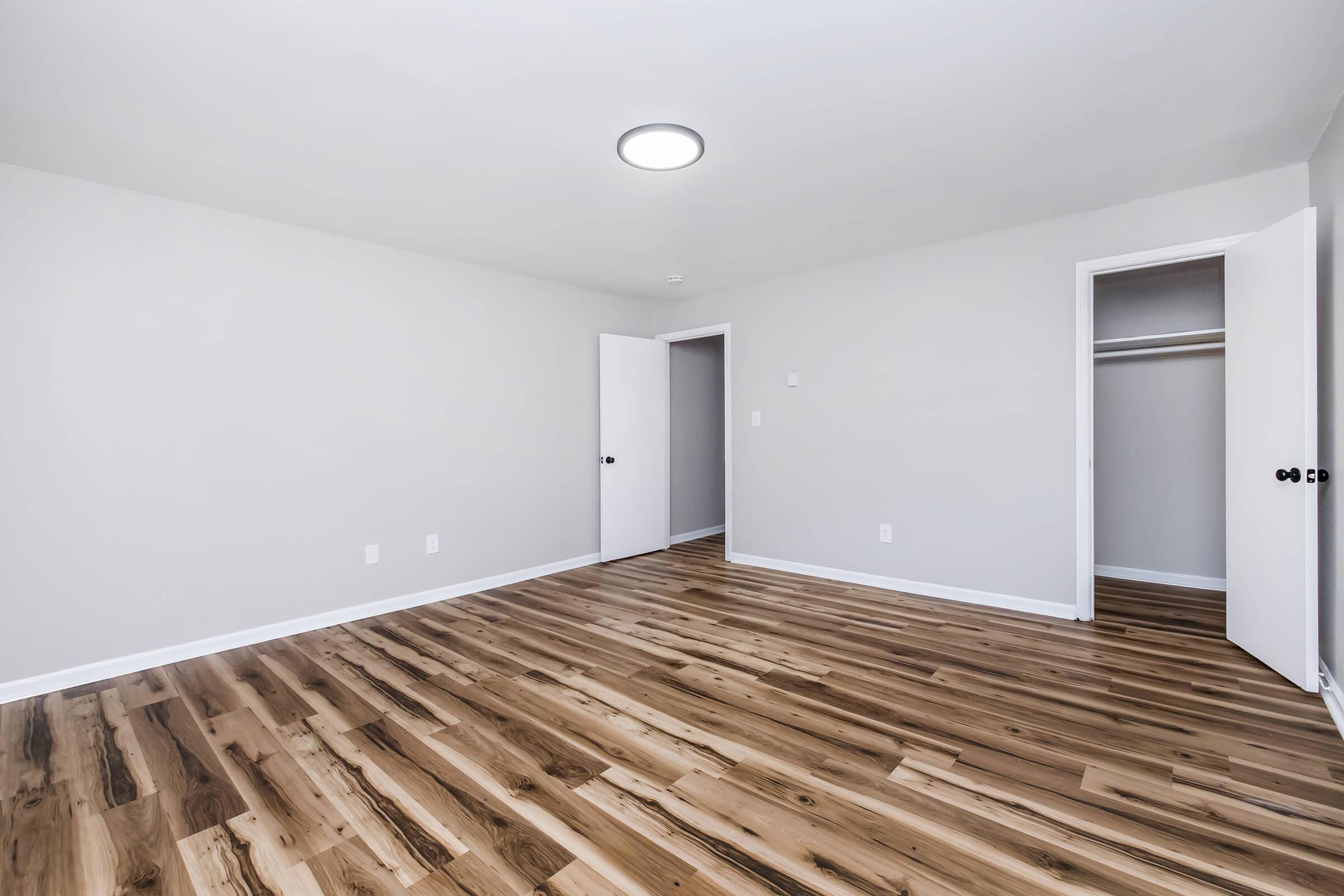 A spacious, empty room with light gray walls and a hardwood floor. It features a simple ceiling light, a closed door on the left, and an open closet with a door on the right. The overall atmosphere is minimalistic and bright, creating a welcoming space ready for furnishing.