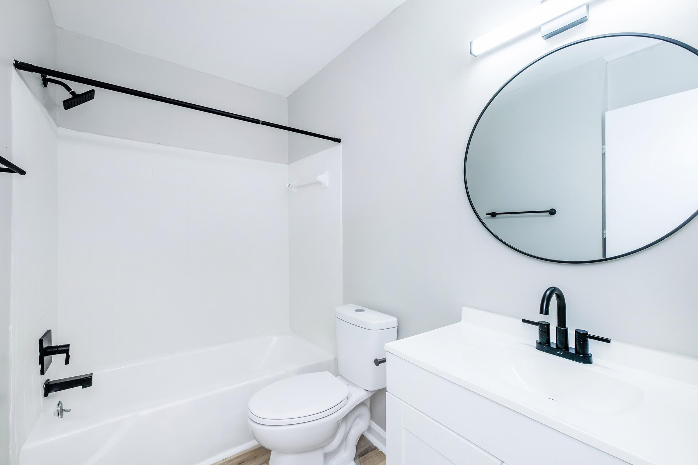 A modern bathroom featuring a white bathtub, toilet, and a sleek vanity with a sink. The walls are painted light gray, complemented by a round mirror and black fixtures. A black towel rack is mounted on the wall, and natural light is provided by a light fixture above the mirror.