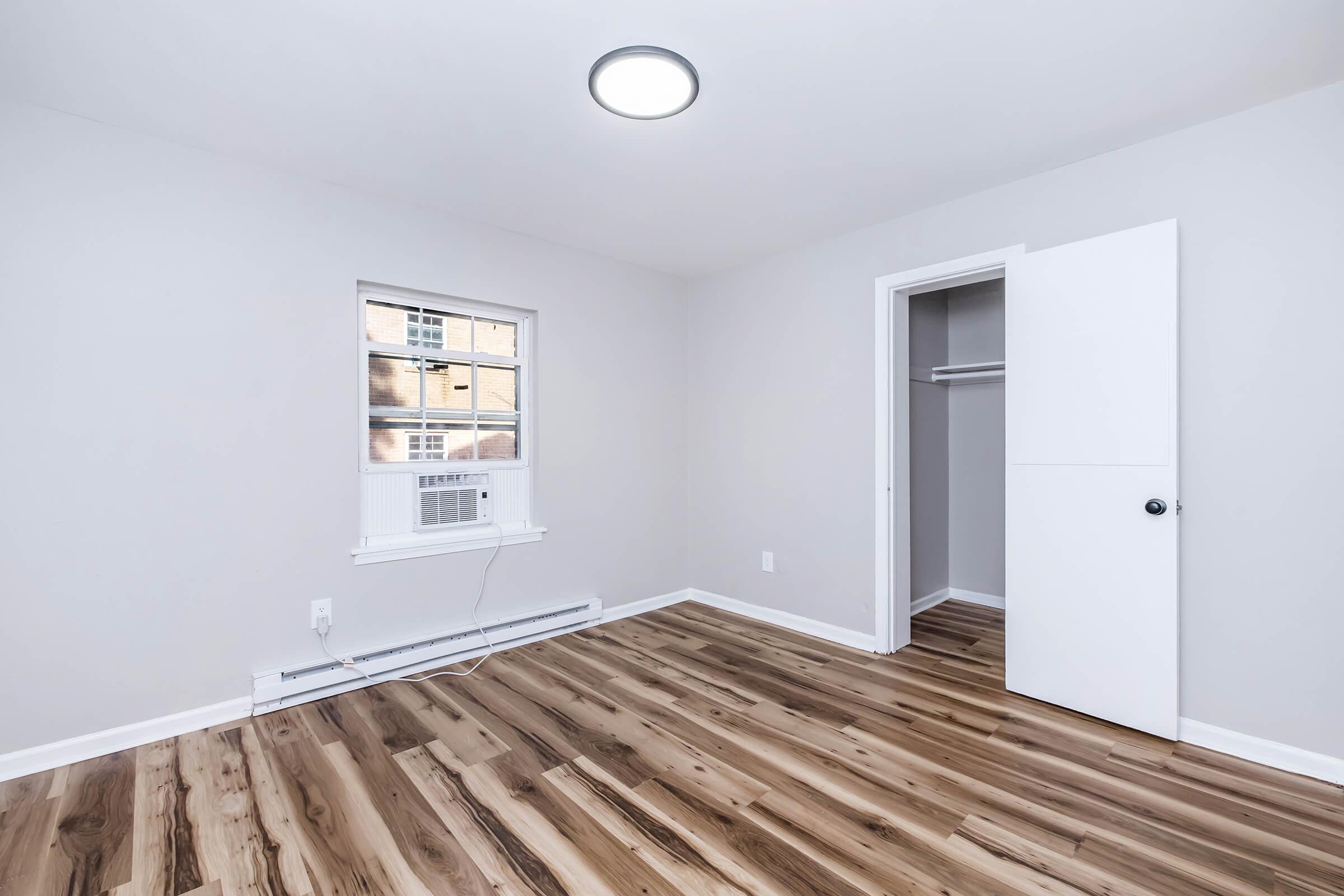 A spacious, empty room with light-colored walls and wooden flooring. It features a window with an air conditioning unit below, and a closet with a white door partially open. The ceiling has a modern circular light fixture, creating a bright and airy atmosphere.