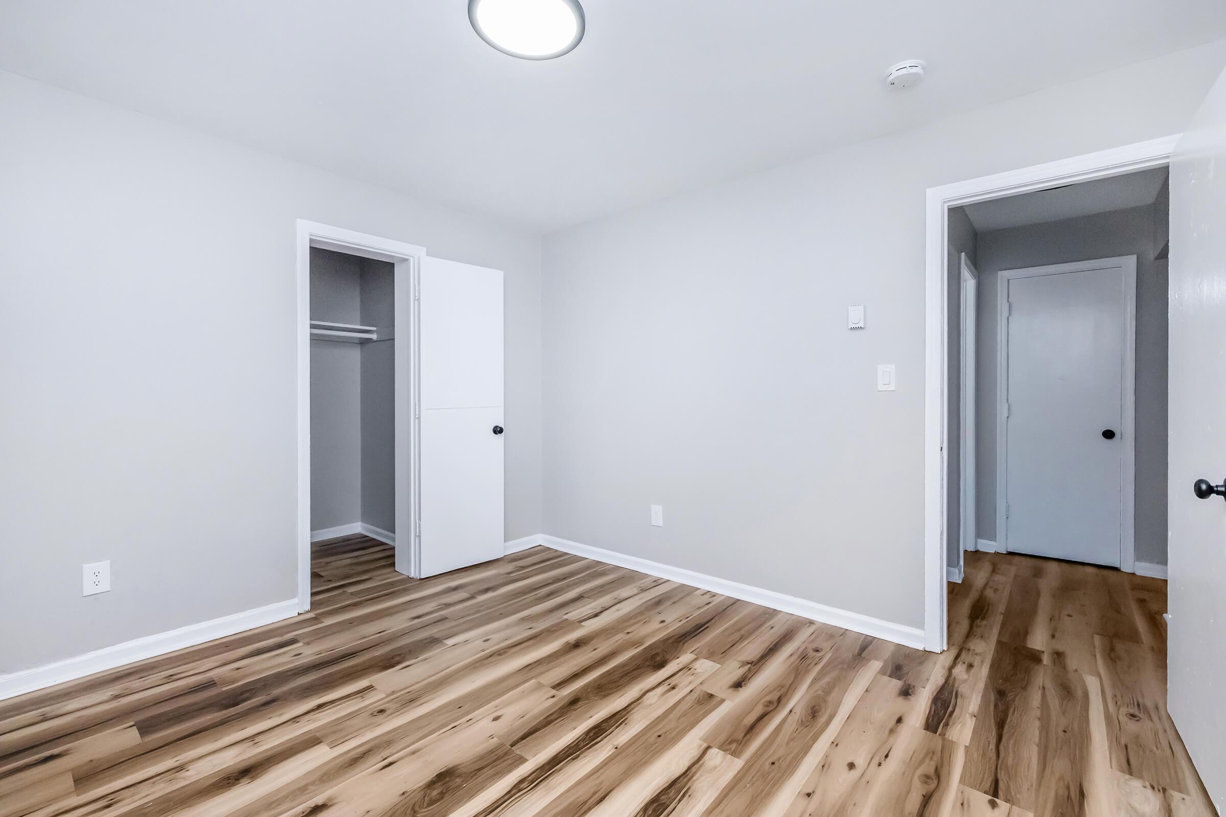 A spacious, empty room with light gray walls and new wooden flooring. It features a closet on one side and two doors leading to adjoining areas. The ceiling has a round light fixture, providing bright illumination, and the overall ambiance is clean and modern.