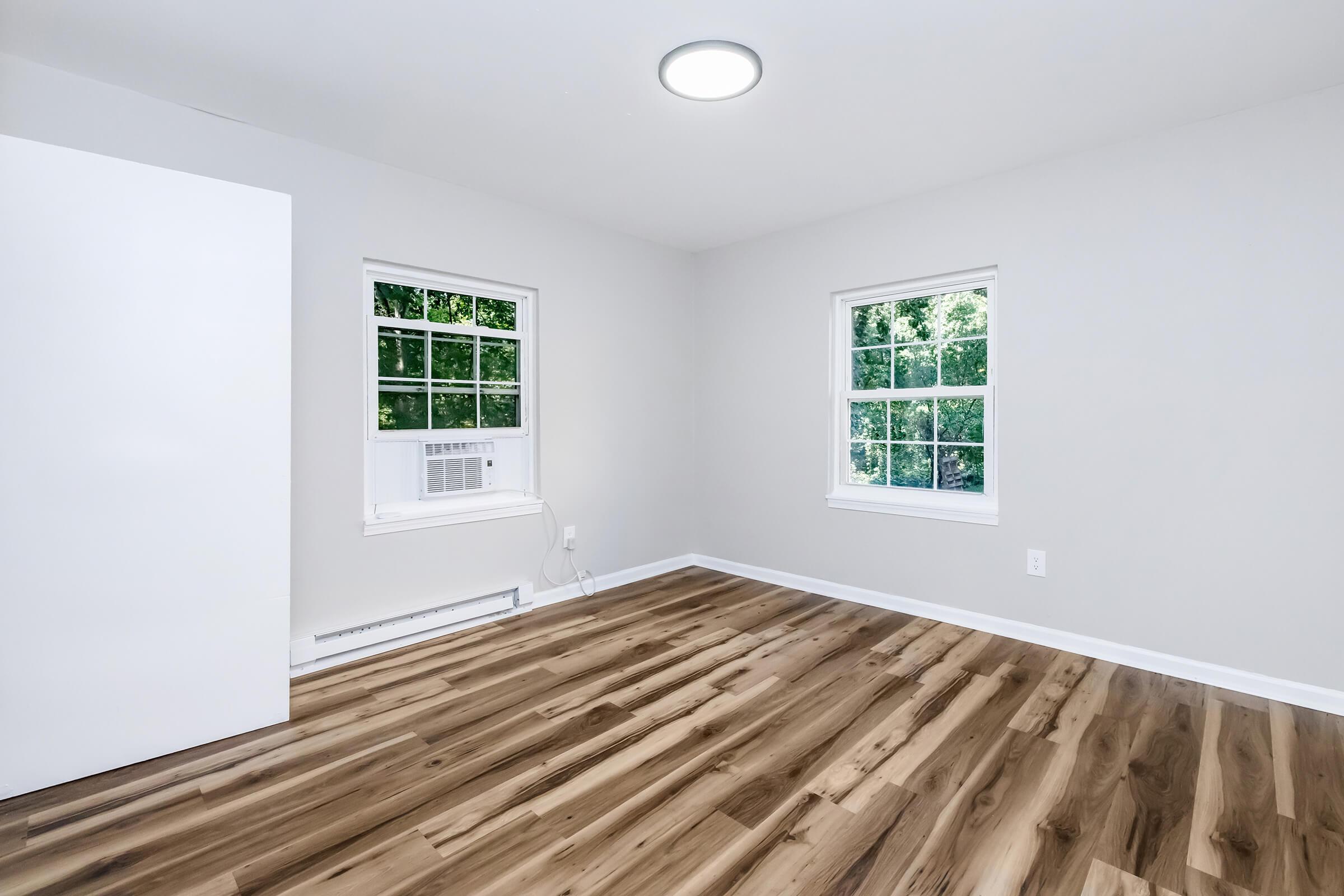 An empty room featuring light gray walls, two windows with white frames, and a white door. The floor is made of wood with a natural finish. An air conditioning unit is installed beneath one of the windows, and there is a ceiling light illuminating the space.