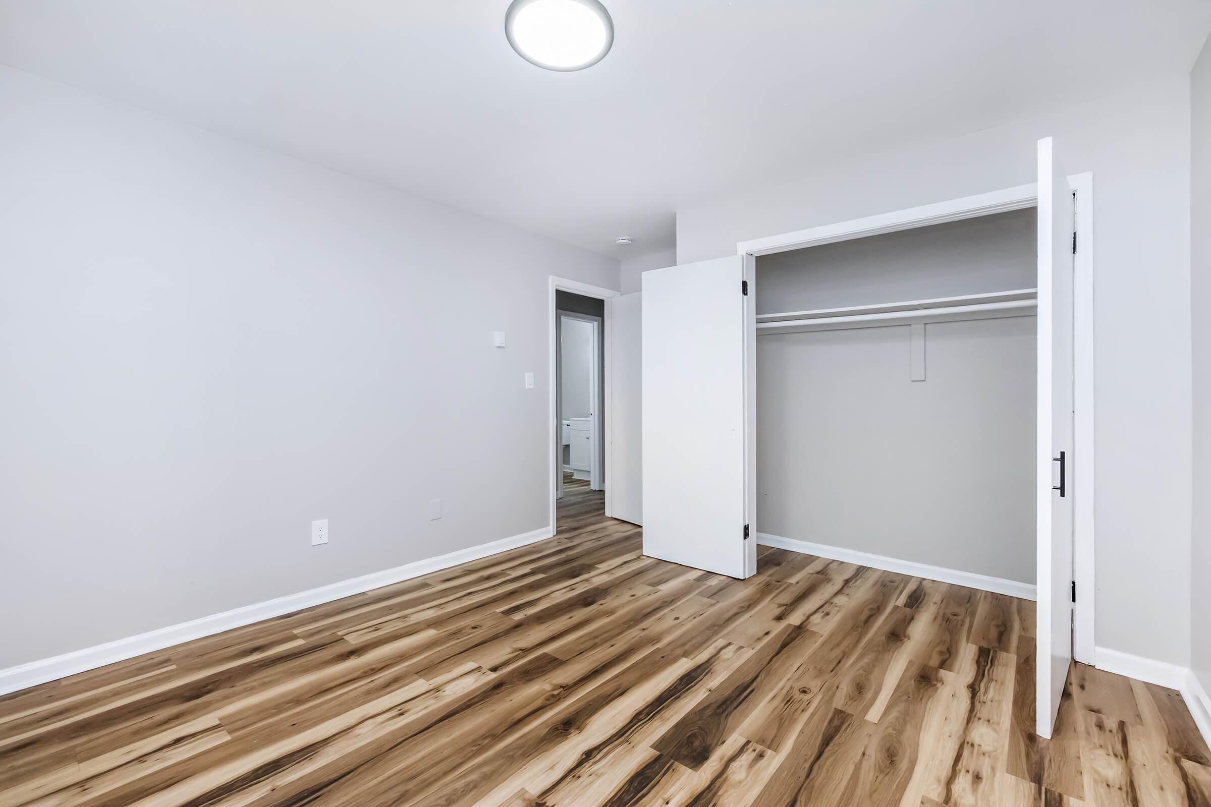 A bright, spacious room with light-colored walls and a medium wood floor. There are two open doors leading to a closet on the left and a hallway. The ceiling features a circular light fixture, adding to the room’s modern and inviting aesthetic. The overall feel is clean and minimalistic.