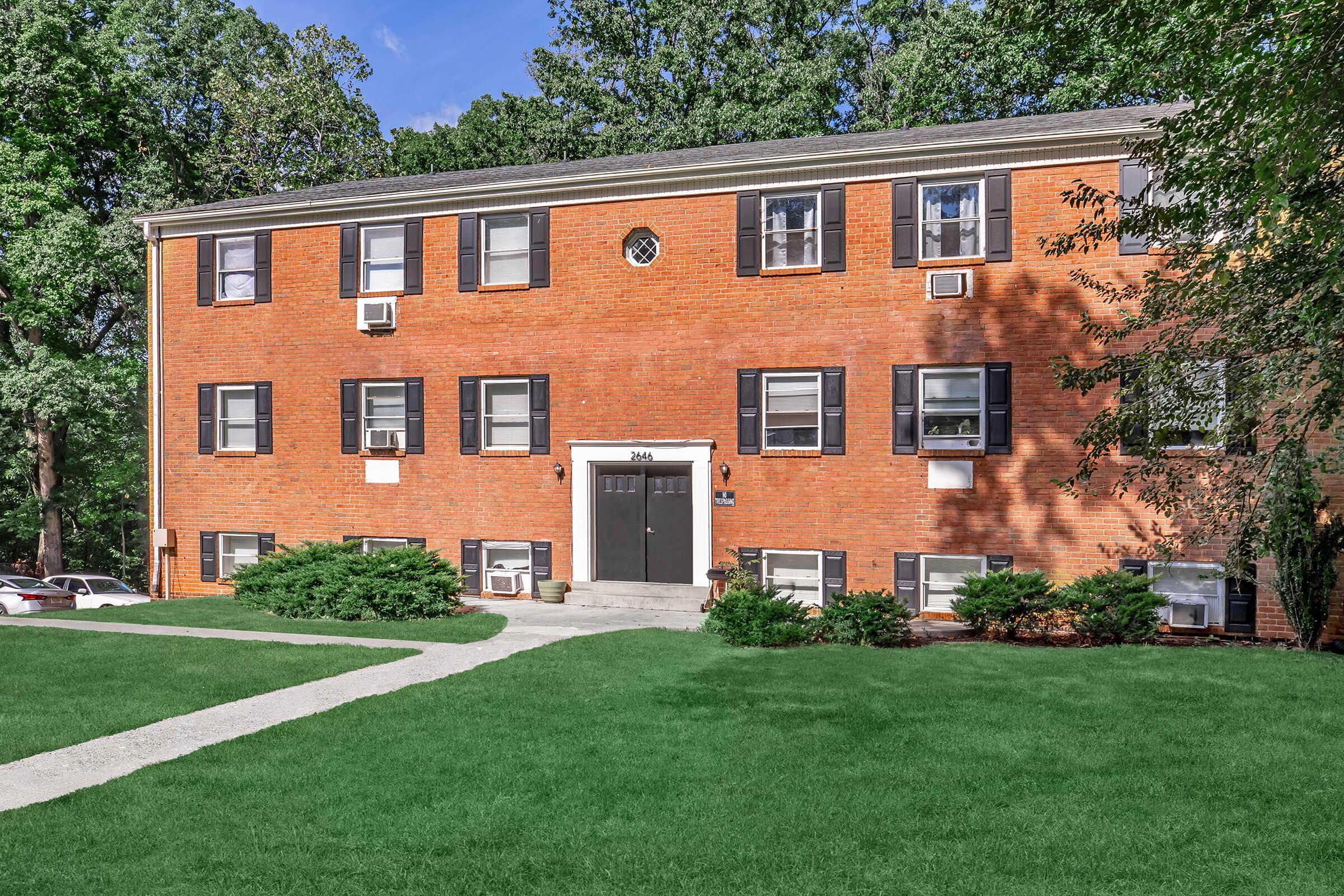 A three-story brick apartment building with a central entrance featuring black doors and shutters. The building is surrounded by green grass and shrubs, with a clear blue sky above. There are multiple air conditioning units visible in the windows, and some parked cars are seen in the background.