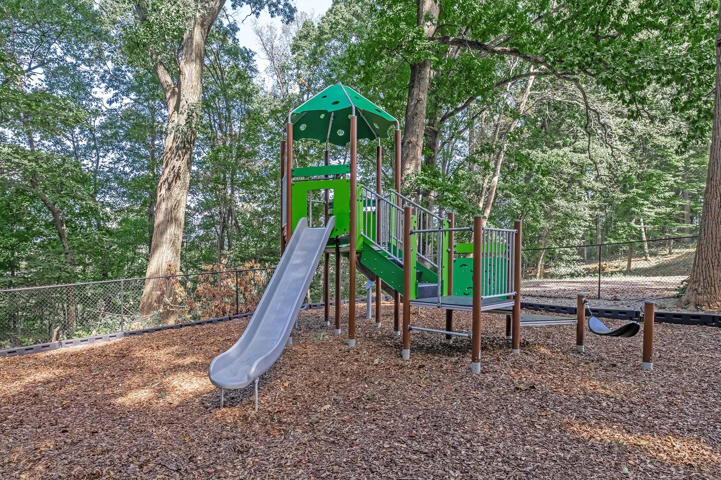 Colorful playground equipment featuring a green play structure with a slide, surrounded by wood chips and trees. The structure includes a climbing area and a railing, set in a natural park-like environment, providing a fun space for children to play.