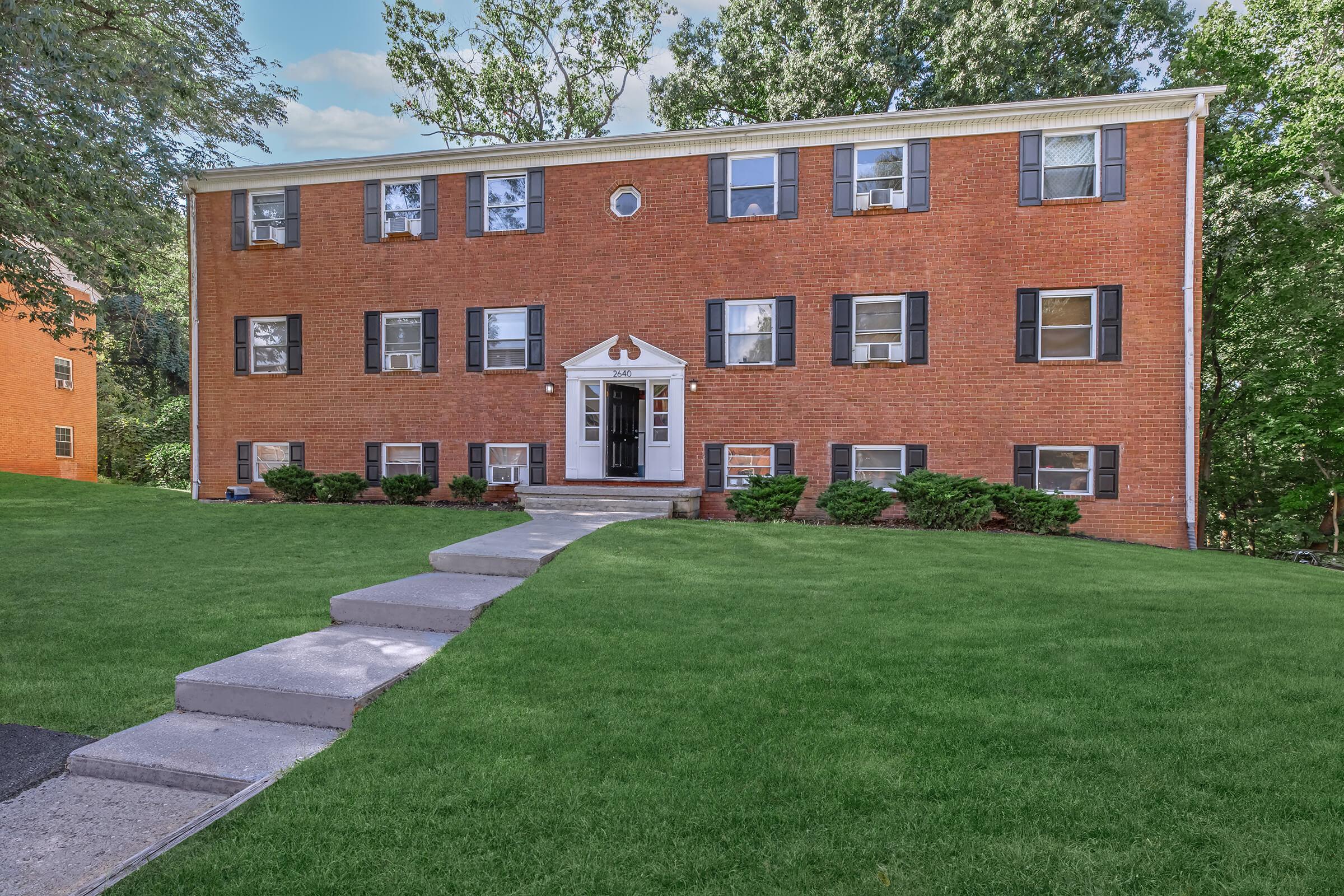 A brick apartment building featuring multiple windows with black shutters, a white front door with a small entrance canopy, surrounded by well-maintained green lawn and shrubs. A paved walkway leads to the entrance, and trees are visible in the background.