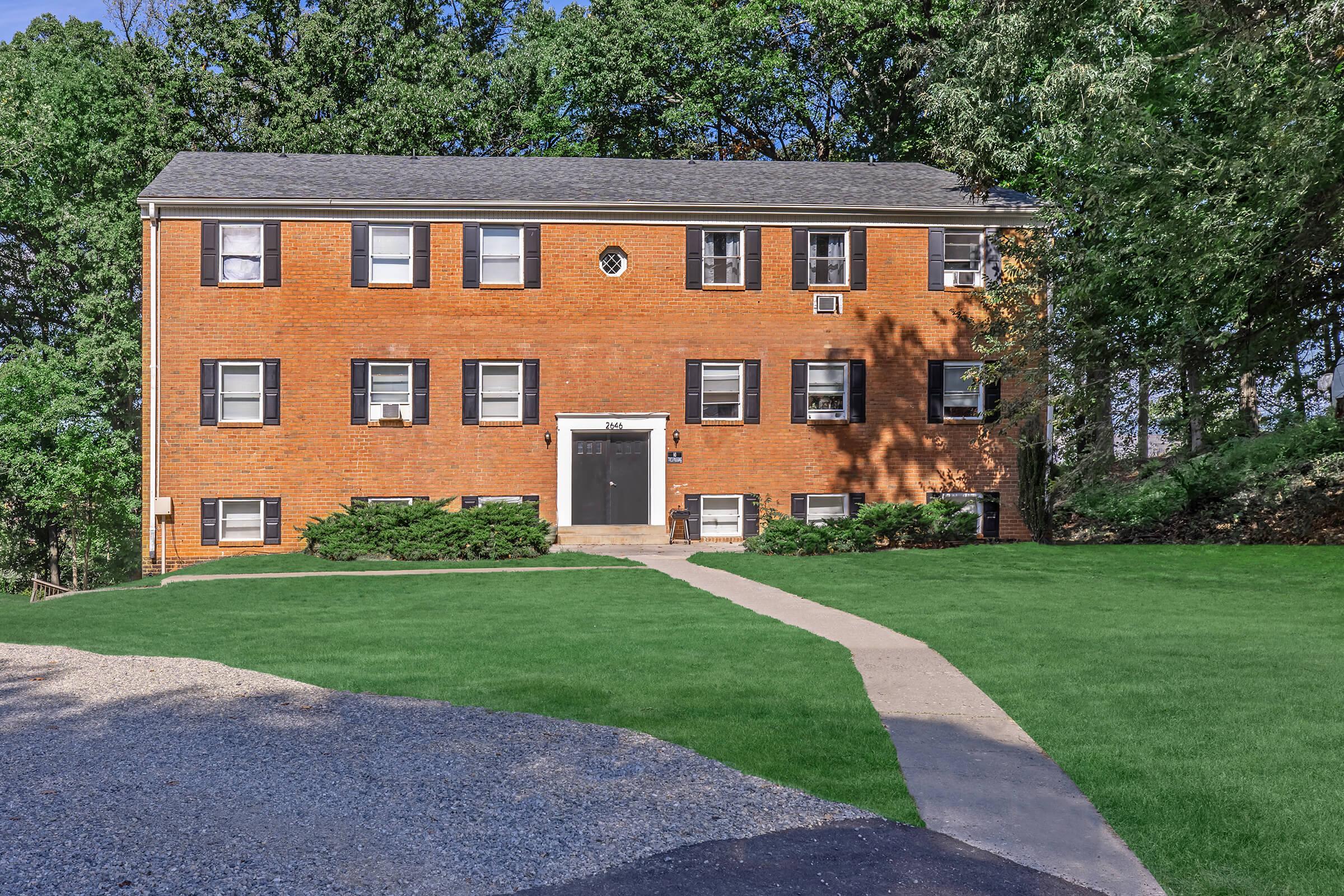 A three-story brick apartment building with a gray roof, featuring multiple windows and a central entrance. The front yard is well-maintained with manicured grass and small shrubs. A paved walkway leads from the driveway to the entrance, surrounded by trees and greenery in the background.
