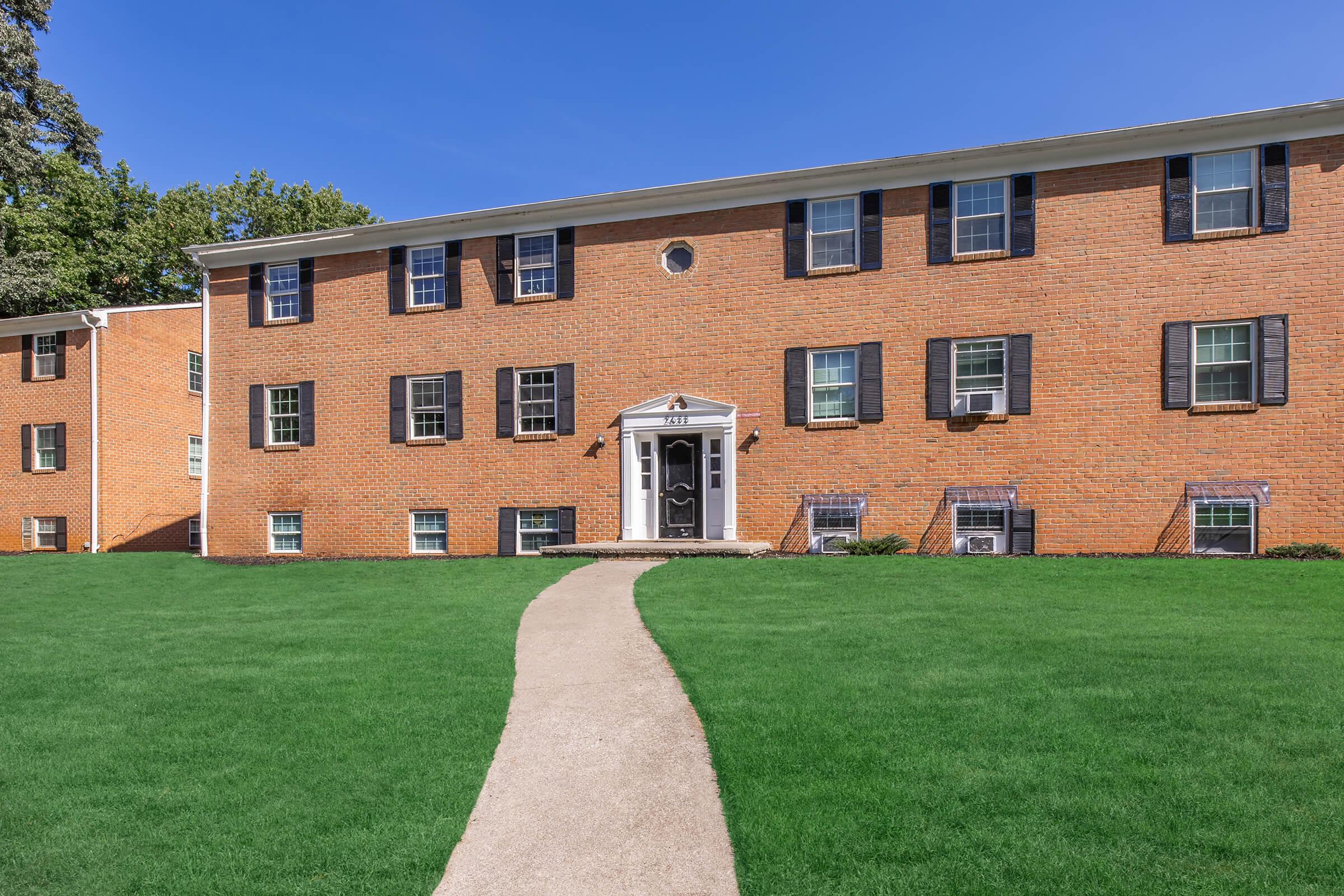 A two-story brick apartment building with multiple windows, black shutters, and a front entrance featuring a white porch. A concrete walkway leads from the entrance to a well-maintained green lawn. The sky is clear and blue, showcasing a bright and sunny day.