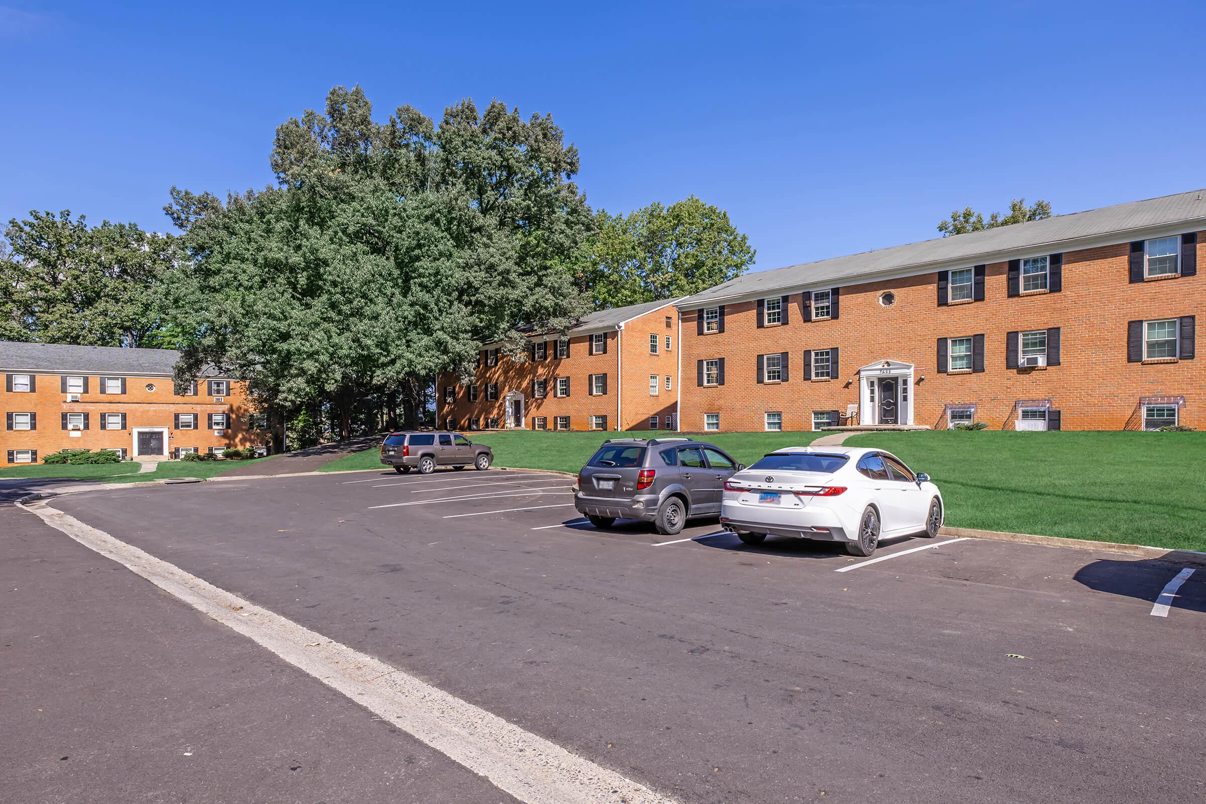 A view of a residential apartment complex featuring two brick buildings and a parking lot with several vehicles. Green trees are visible in the background, and the sky is clear and blue. The grassy area surrounding the buildings adds to the pleasant, suburban atmosphere.