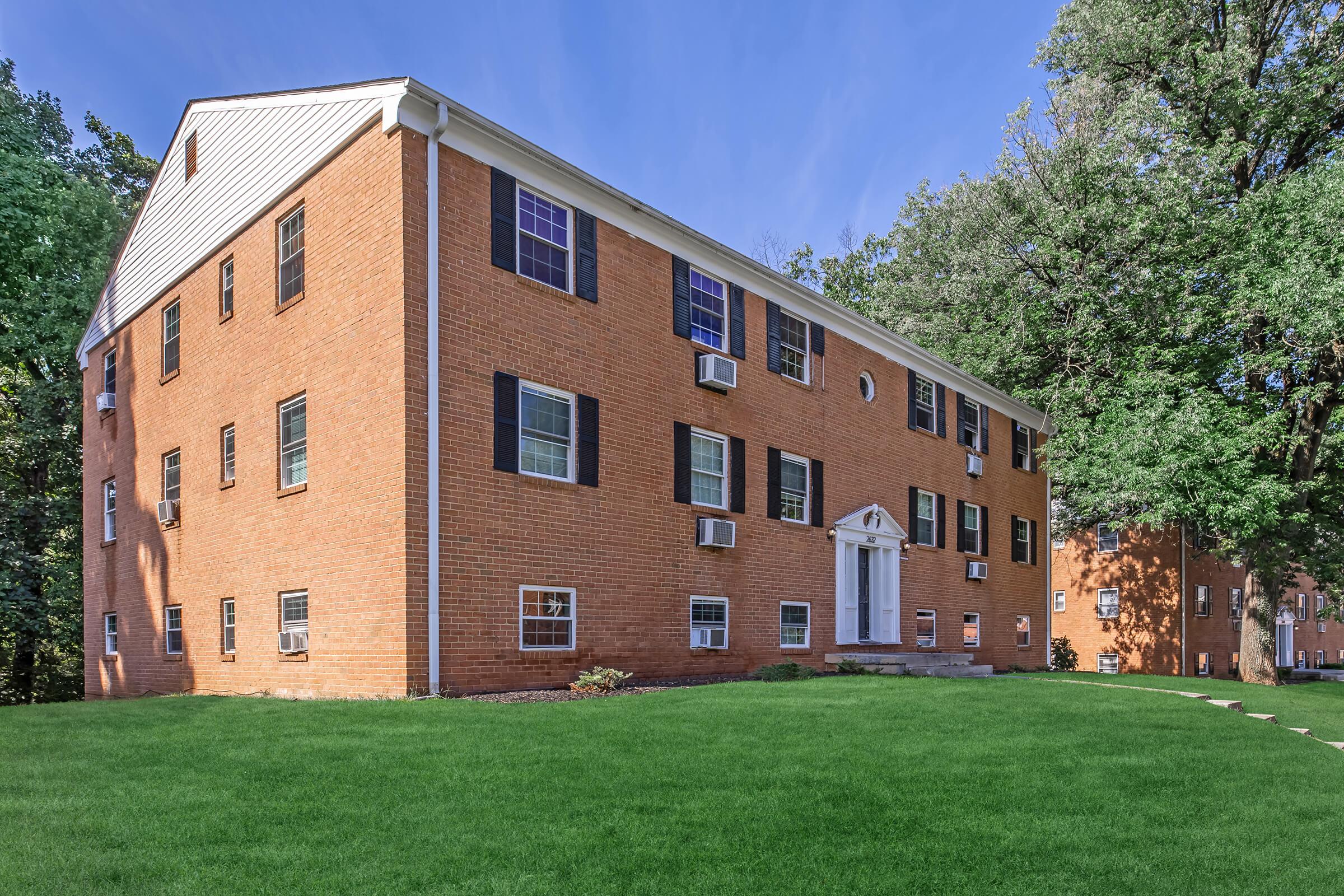 A brick apartment building with multiple windows and air conditioning units, set on a green lawn. The building features white accents and a central entrance, surrounded by trees. The sky is clear with a hint of blue, creating a tranquil suburban atmosphere.