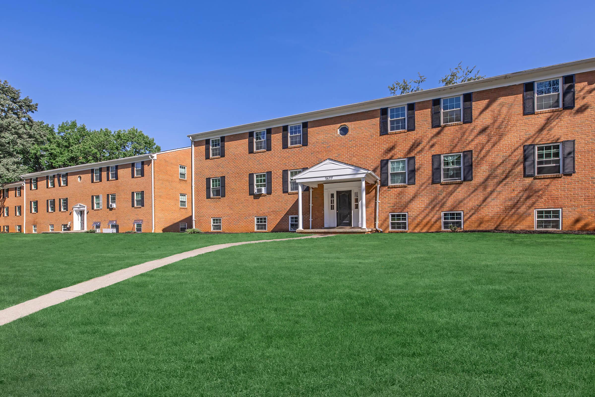A row of brick apartment buildings with white trim, set against a clear blue sky. The lawn in front is green and well-maintained, with a paved walkway leading to the entrance of the nearest building. Trees are visible in the background, adding to the suburban atmosphere.