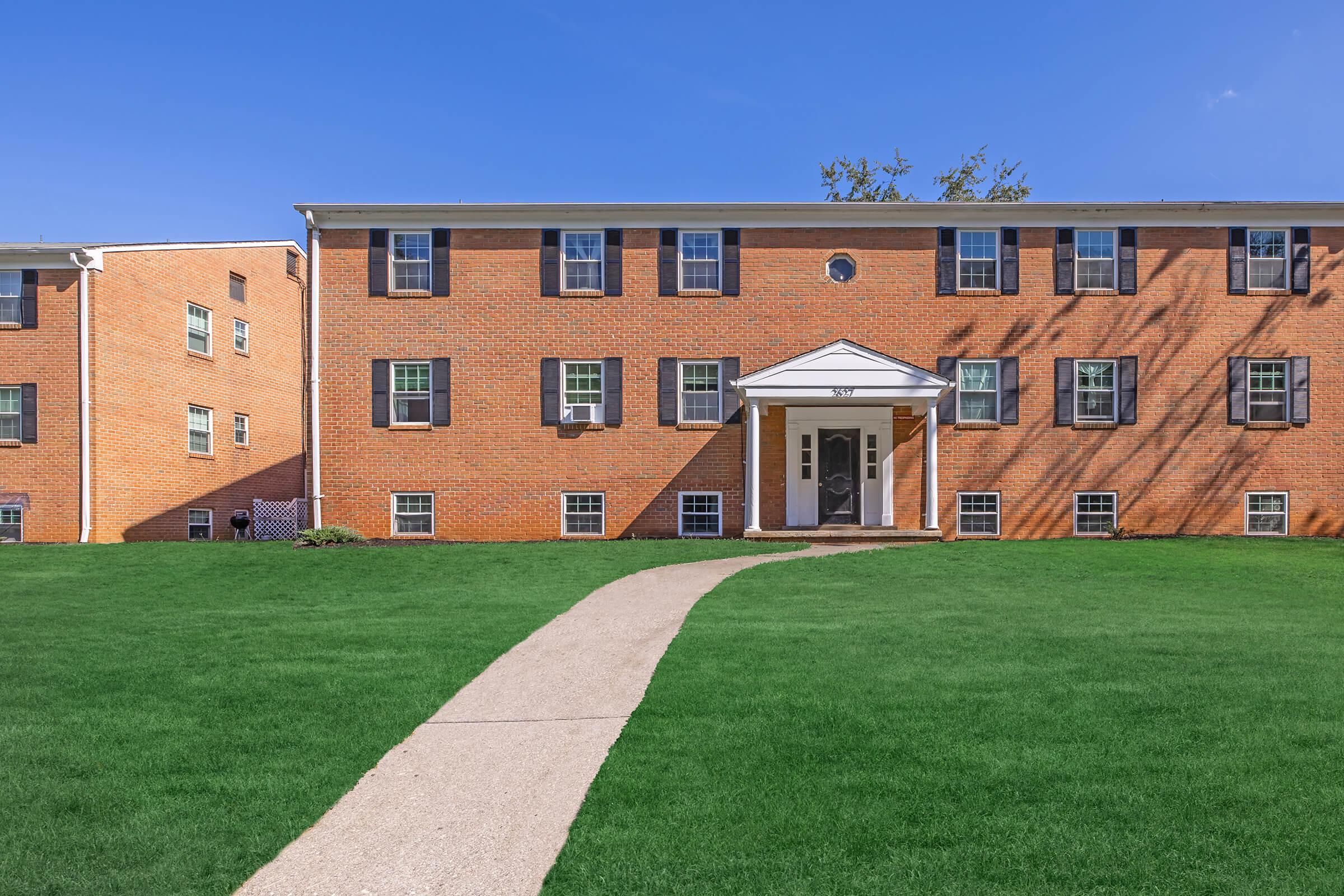 A two-story brick apartment building with a central entrance, flanked by windows, set against a clear blue sky. A well-maintained lawn and a concrete path lead to the entrance, highlighting a tidy exterior. The surrounding area is open and grassy, suggesting a residential neighborhood.