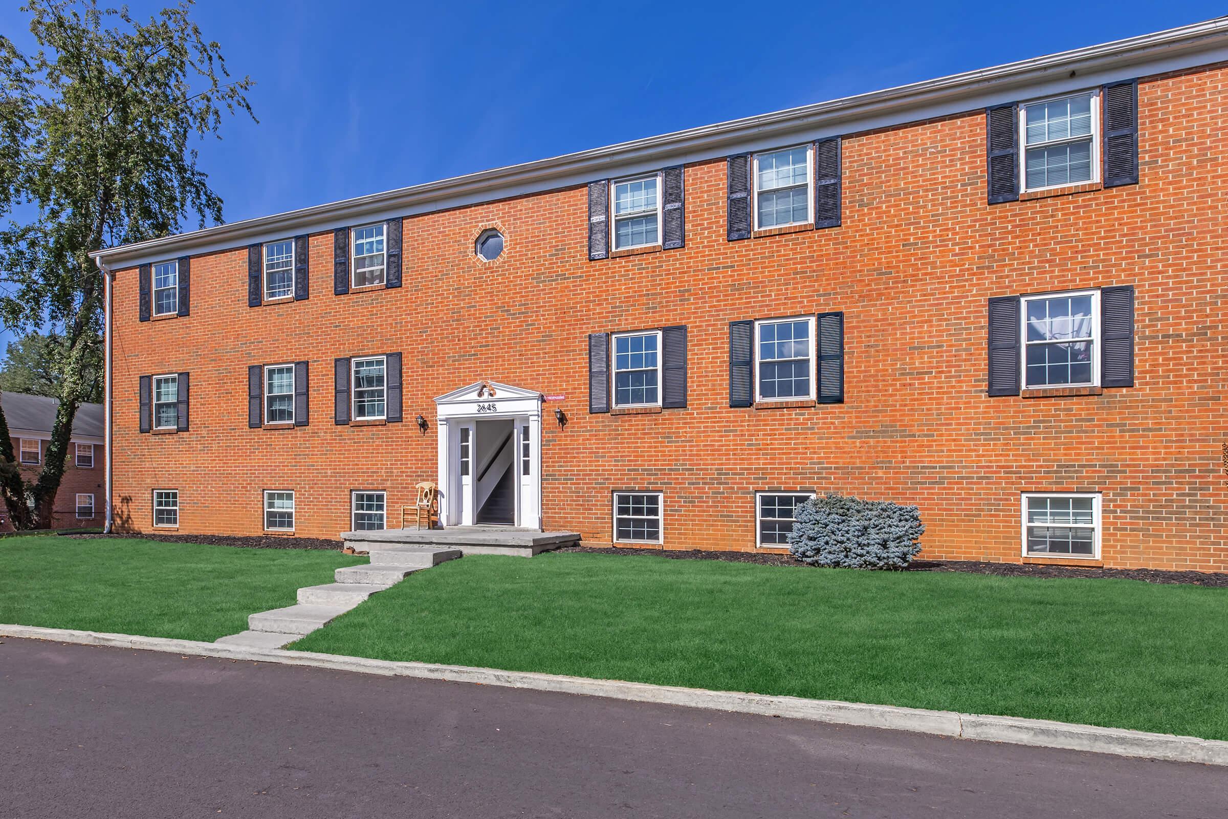 A brick apartment building with multiple windows, a central entrance featuring white doors and a small porch, surrounded by well-maintained grass. The sky is clear blue, and there are trees and shrubs nearby. The driveway is paved, leading up to the entrance.