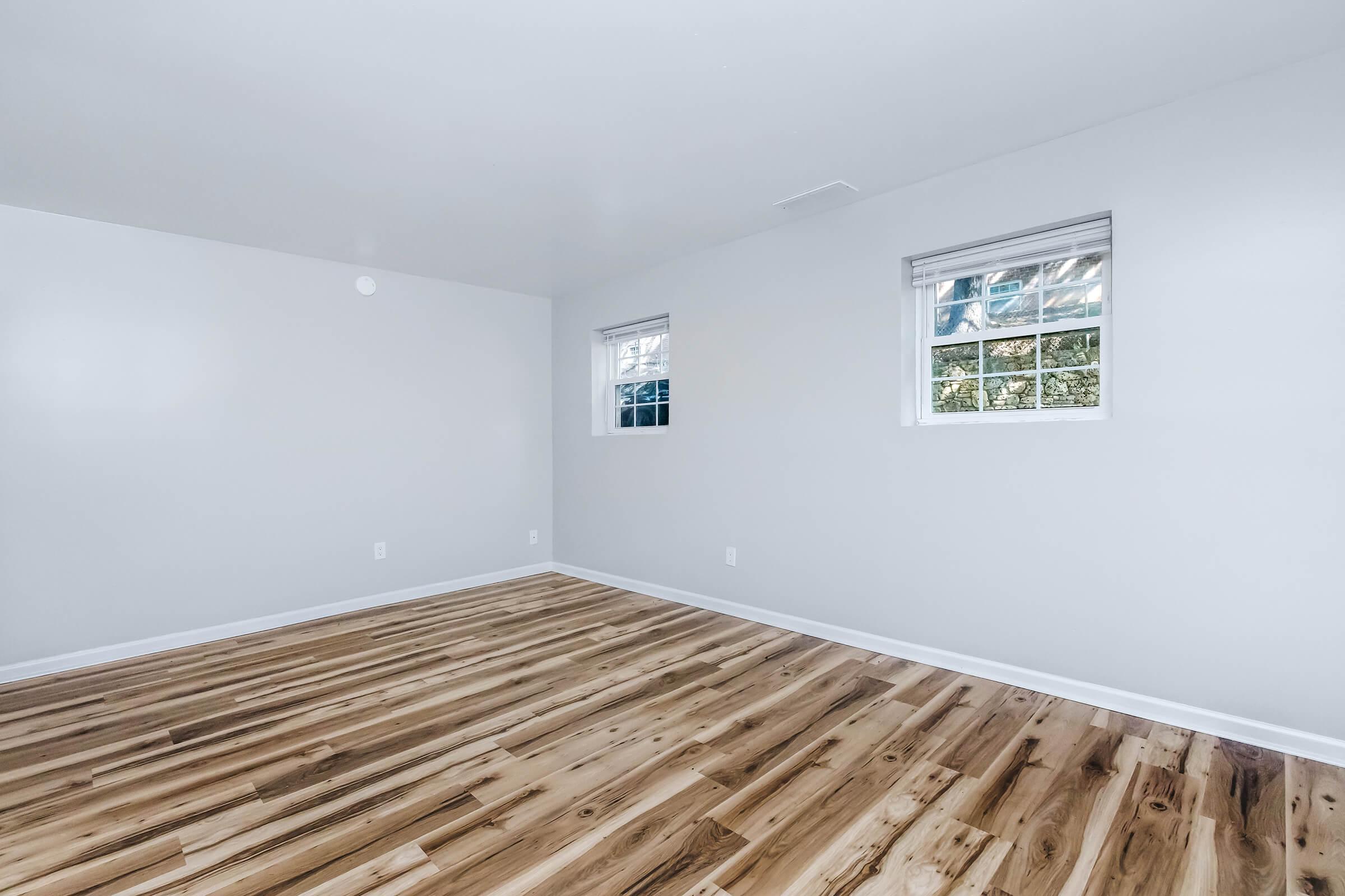 Empty room with light-colored walls and polished wood flooring. Two windows provide natural light, and there are no furnishings or decorations visible. The space appears clean and well-maintained, suitable for various uses.