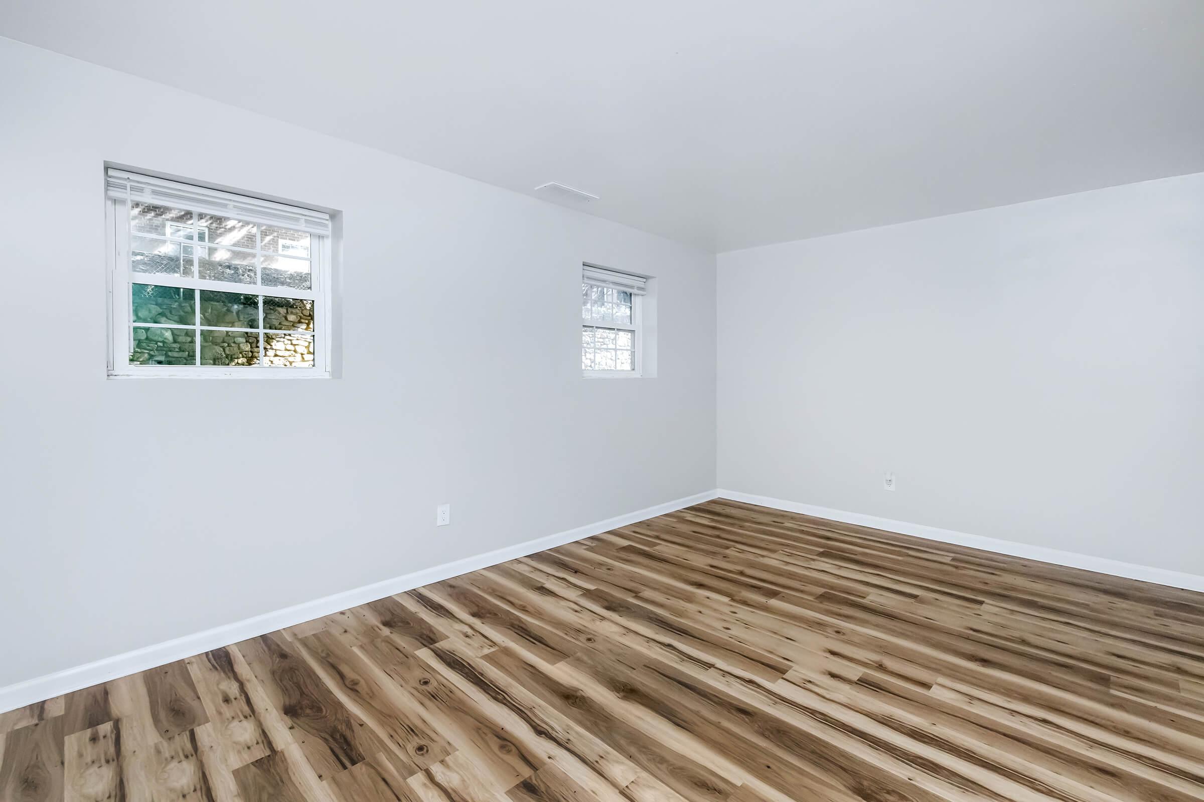 A clean, empty room featuring light-colored walls, two small windows with white trim, and a hardwood floor with a natural wood finish. The space is well-lit and minimalistic, ideal for various uses or as a blank canvas for decorating.