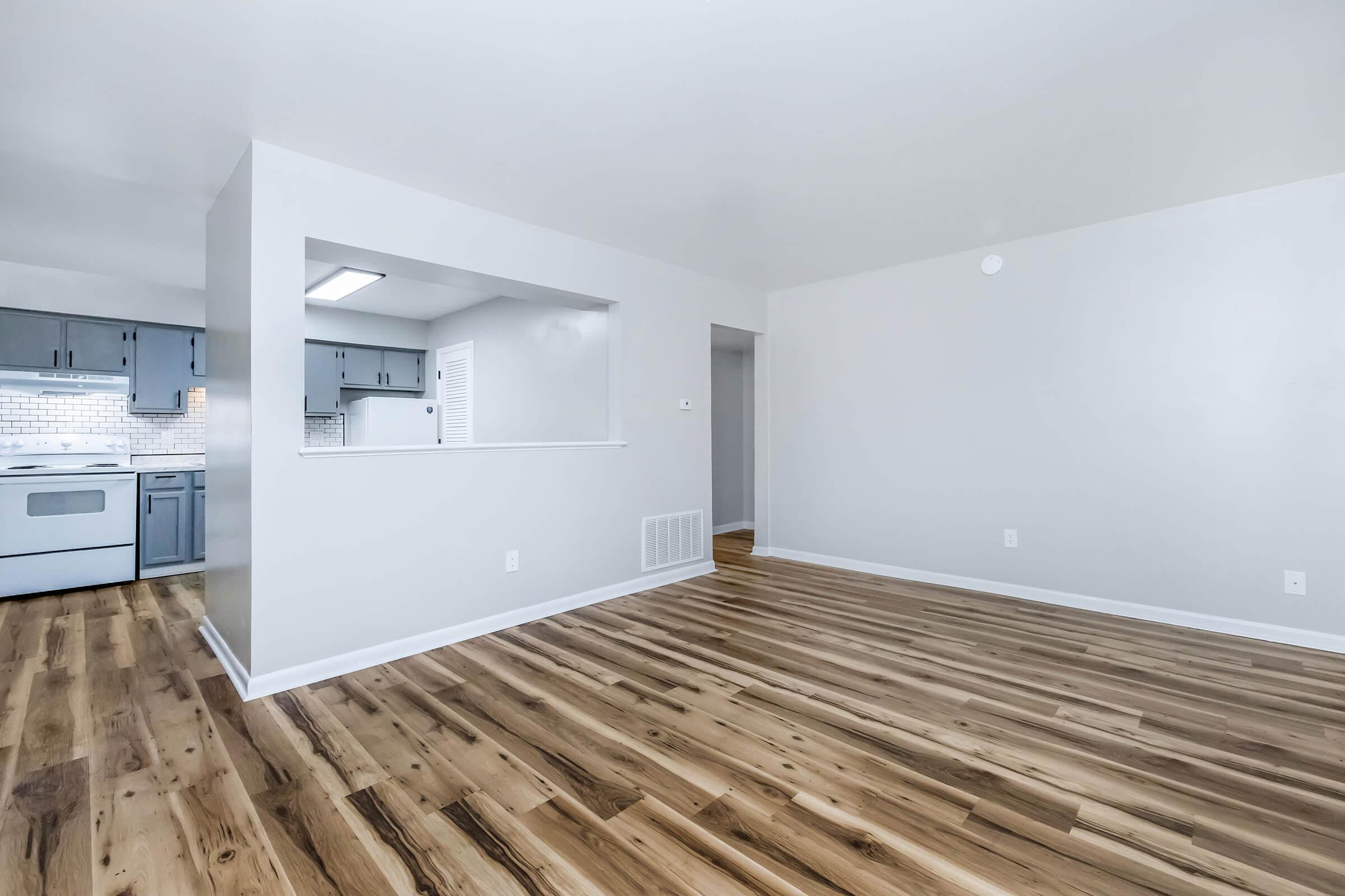 Spacious, modern apartment interior featuring light-colored walls, hardwood flooring, and an open layout. A windowed kitchen is partially visible, showcasing cabinetry and appliances. The room is well-lit and inviting, perfect for a comfortable living space.
